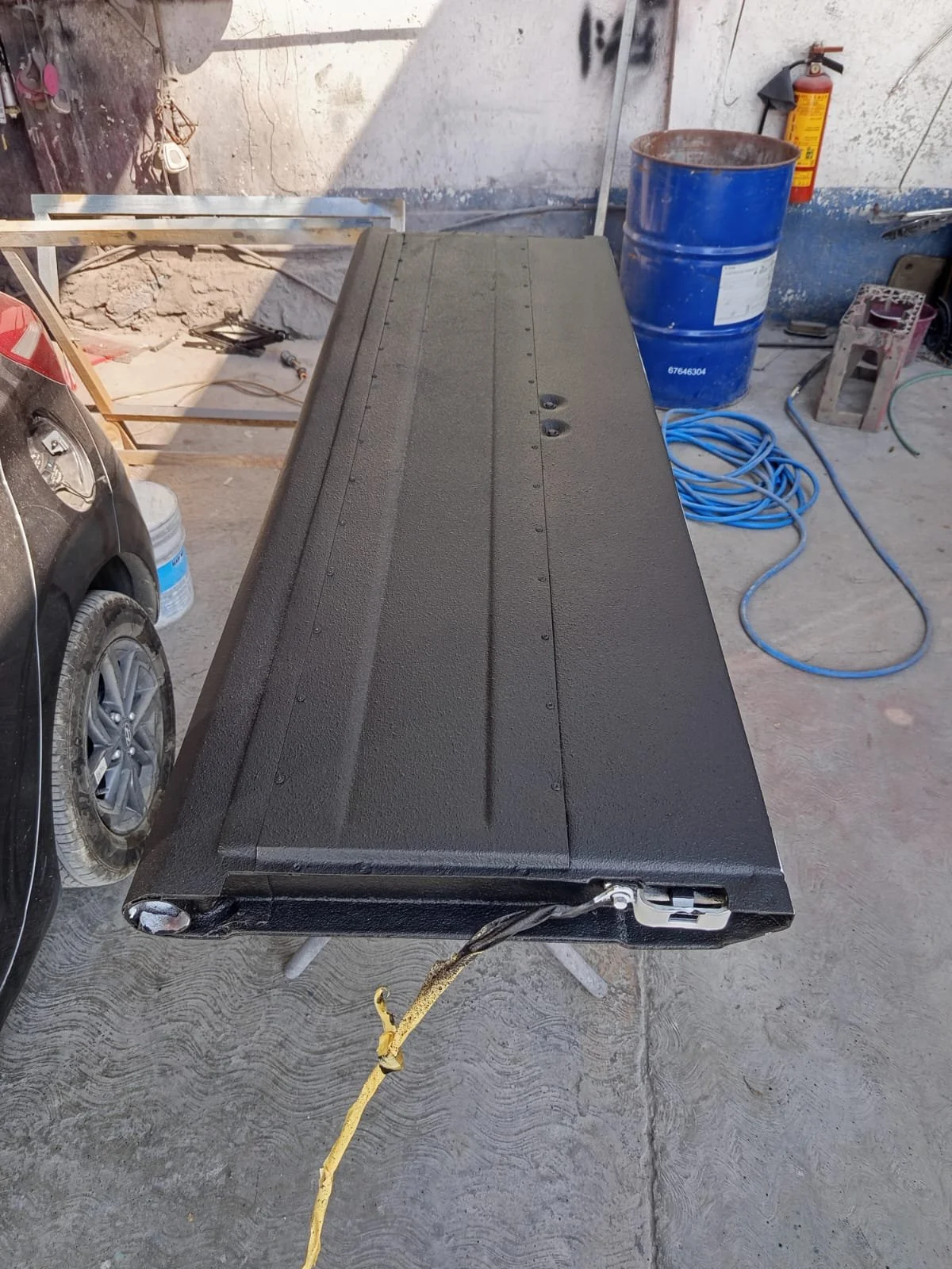 Mesabanco negro en un taller de trabajo, junto a una manguera azul y un bate de metal con pintura azul, en un espacio con paredes en estado de desgaste.