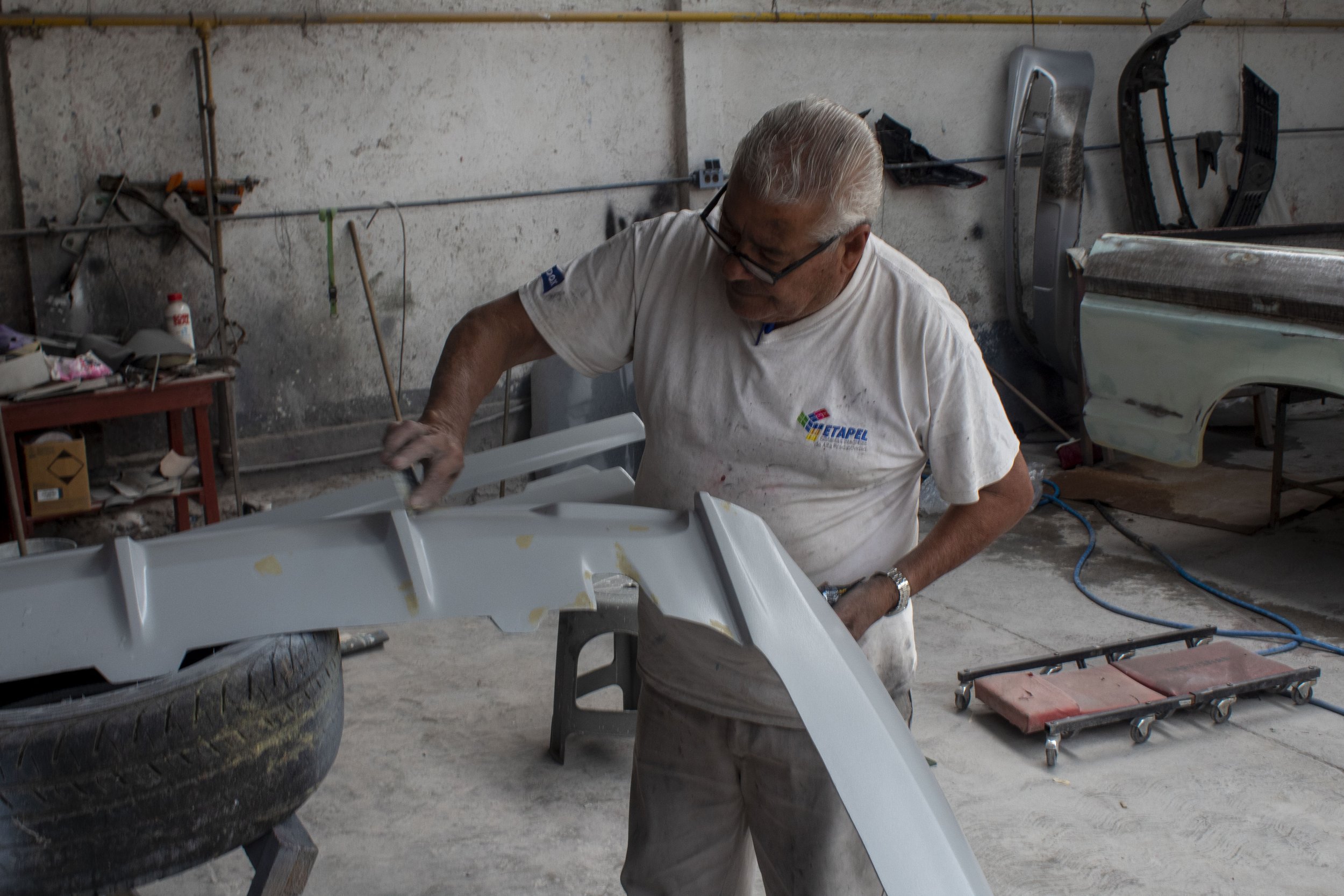 Hombre mayor trabajando en un taller de carrocería, pintando una parte de carro de color gris, rodeado de herramientas y piezas de automóvil.