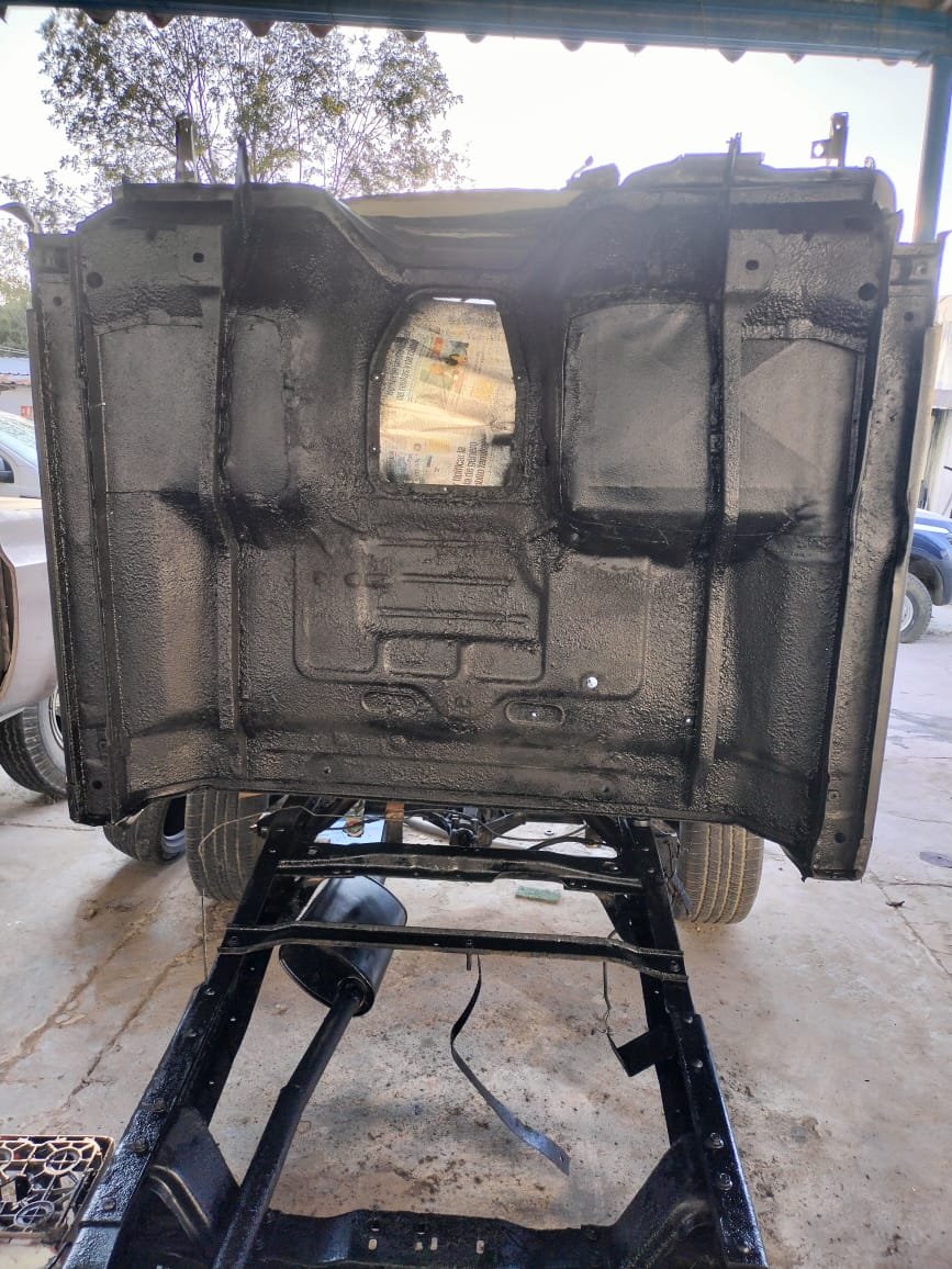 Chasis de vehículo montado en un soporte, visto desde abajo, con pintura negra. Fondo con árboles y autos estacionados.