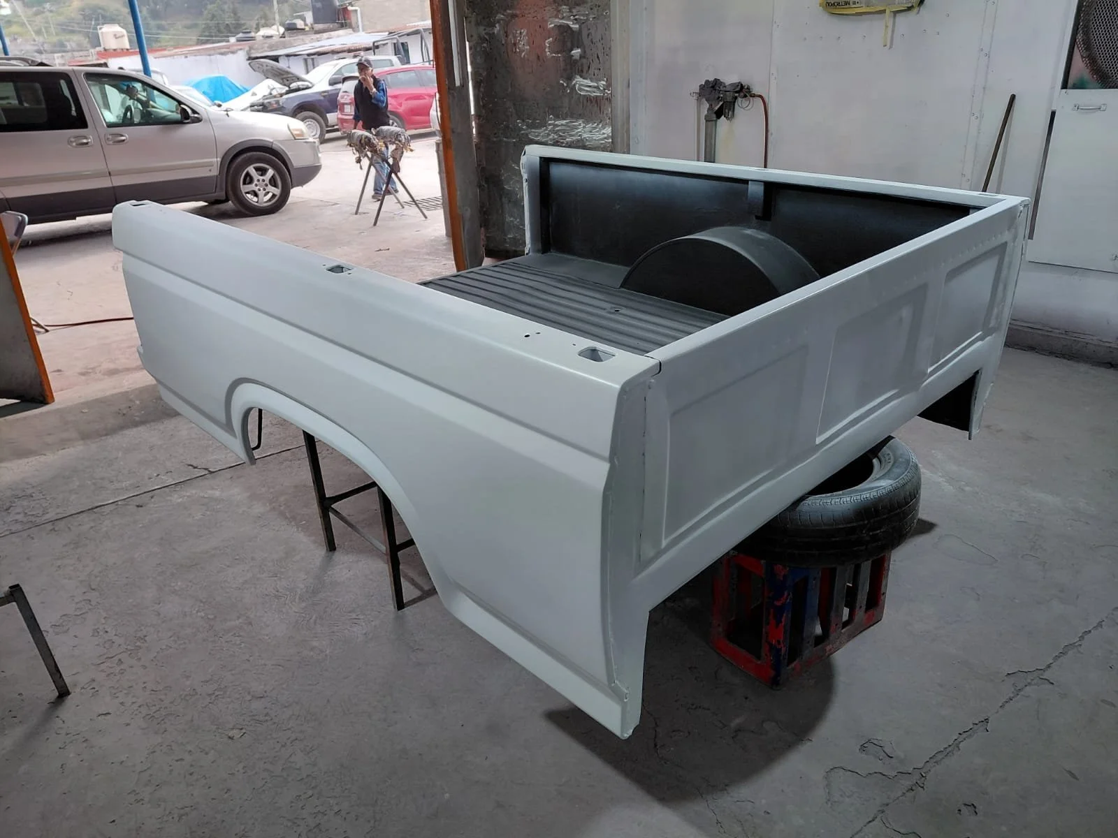 Cabina de camion en proceso de pintado en taller, con varios coches estacionados afuera y una persona trabajando en el fondo.