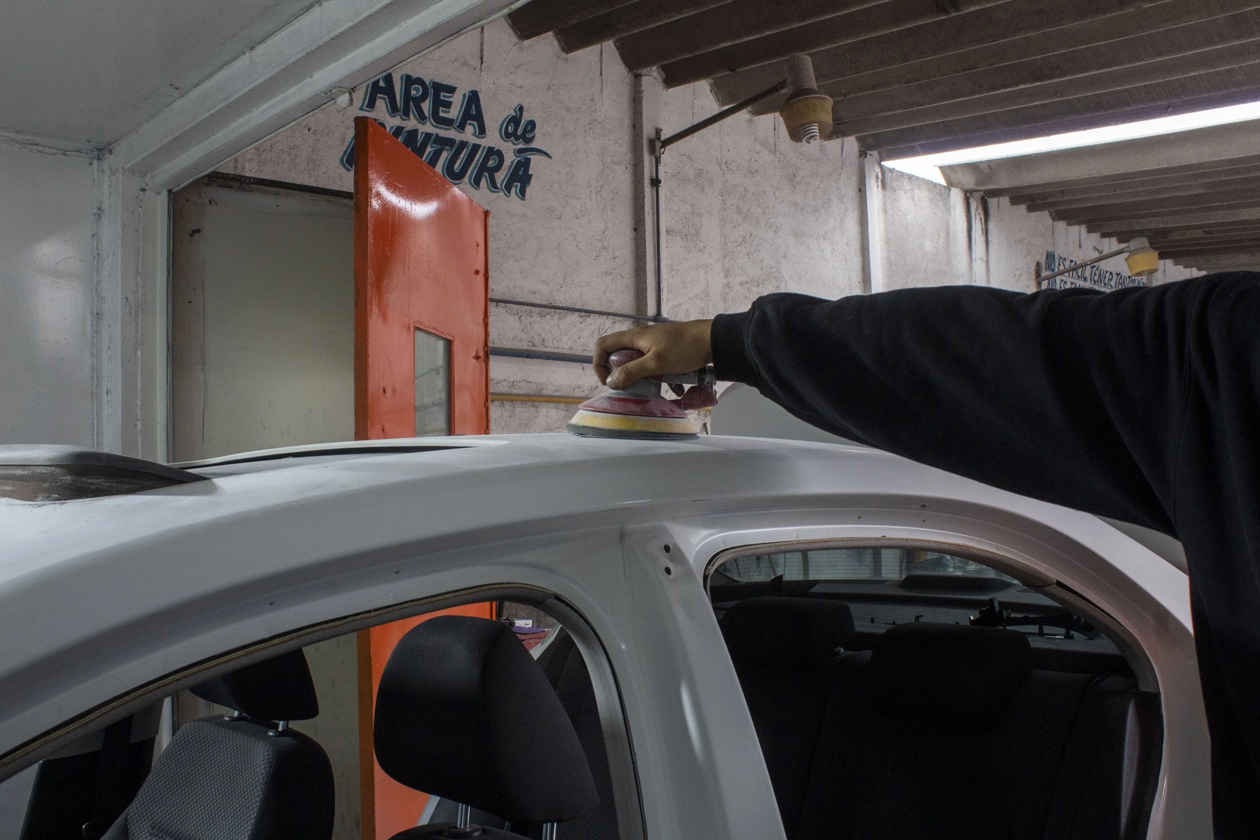 Persona usando una pulidora en un coche en una área de taller o lavado de autos, con señales y estructuras de fondo.