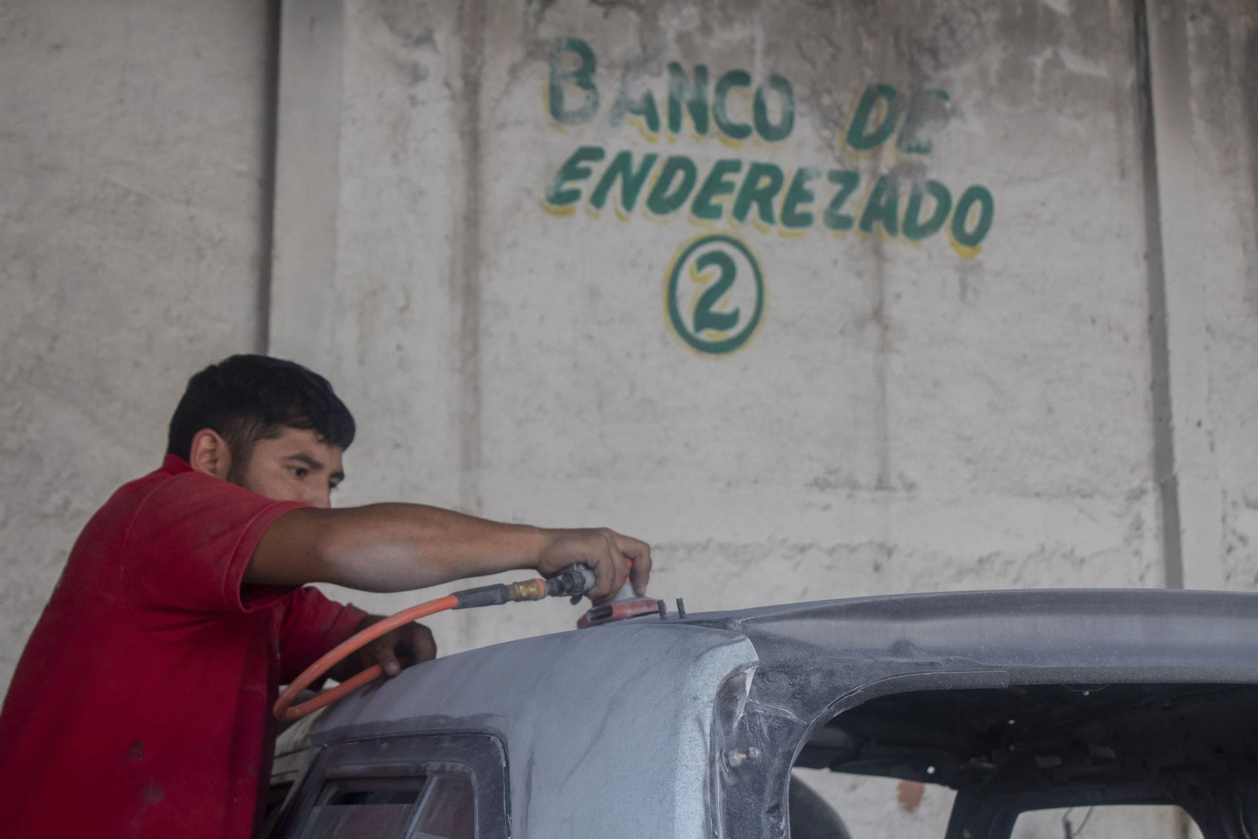 Un joven trabajando en un coche en un taller, usando una herramienta y vestido con una chaqueta roja. En la pared hay un cartel que dice 'Banco de Enderezado' con un número 2.