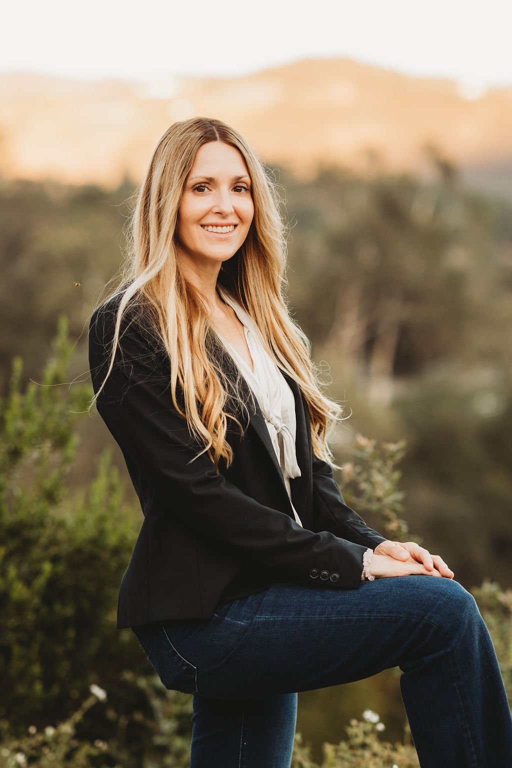 A smiling woman with long blonde hair sitting outdoors with a blurred natural background, wearing a black blazer, white blouse, and blue jeans.