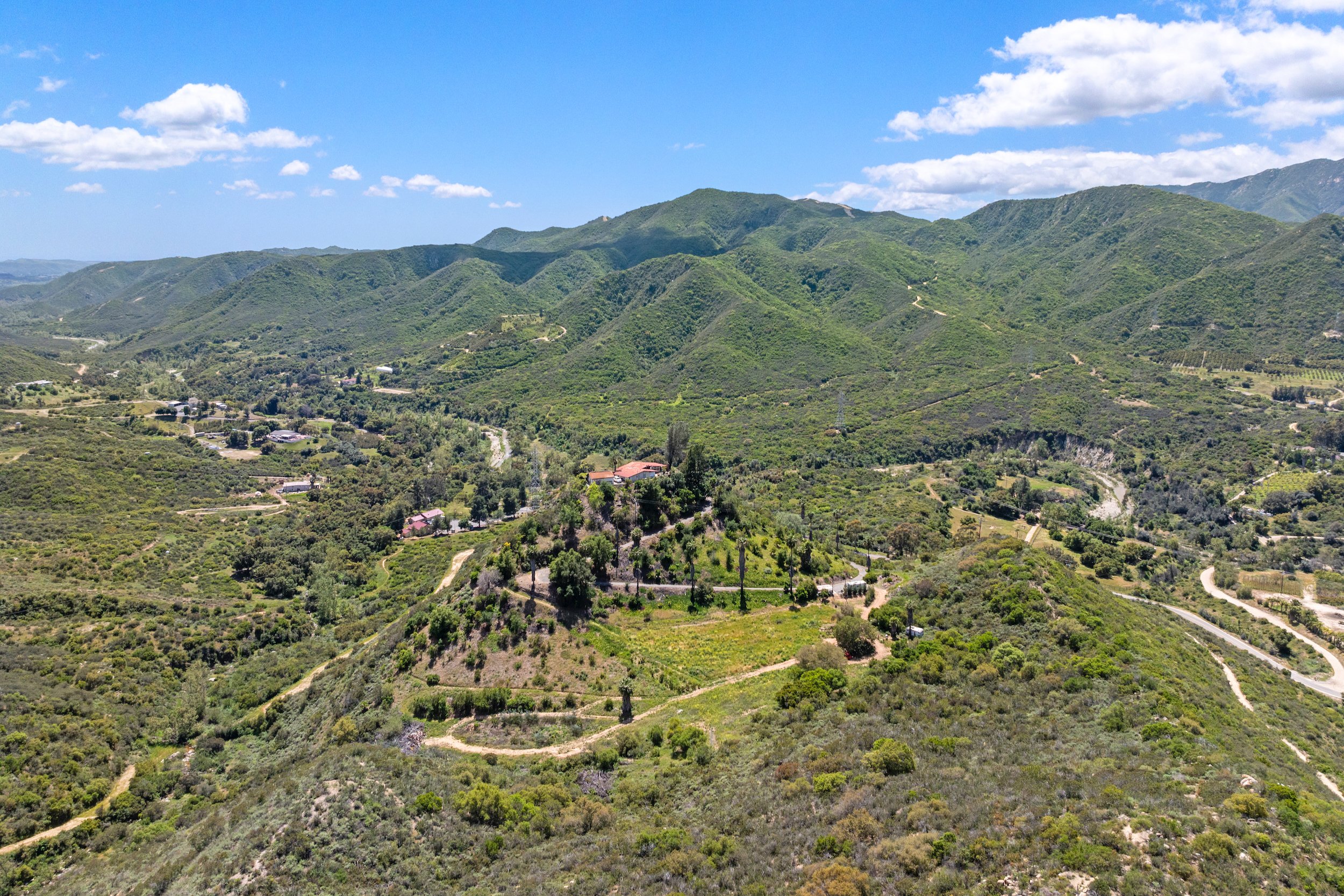 A scenic view of a lush, green mountain range with winding dirt roads and sparse buildings under a partly cloudy blue sky.
