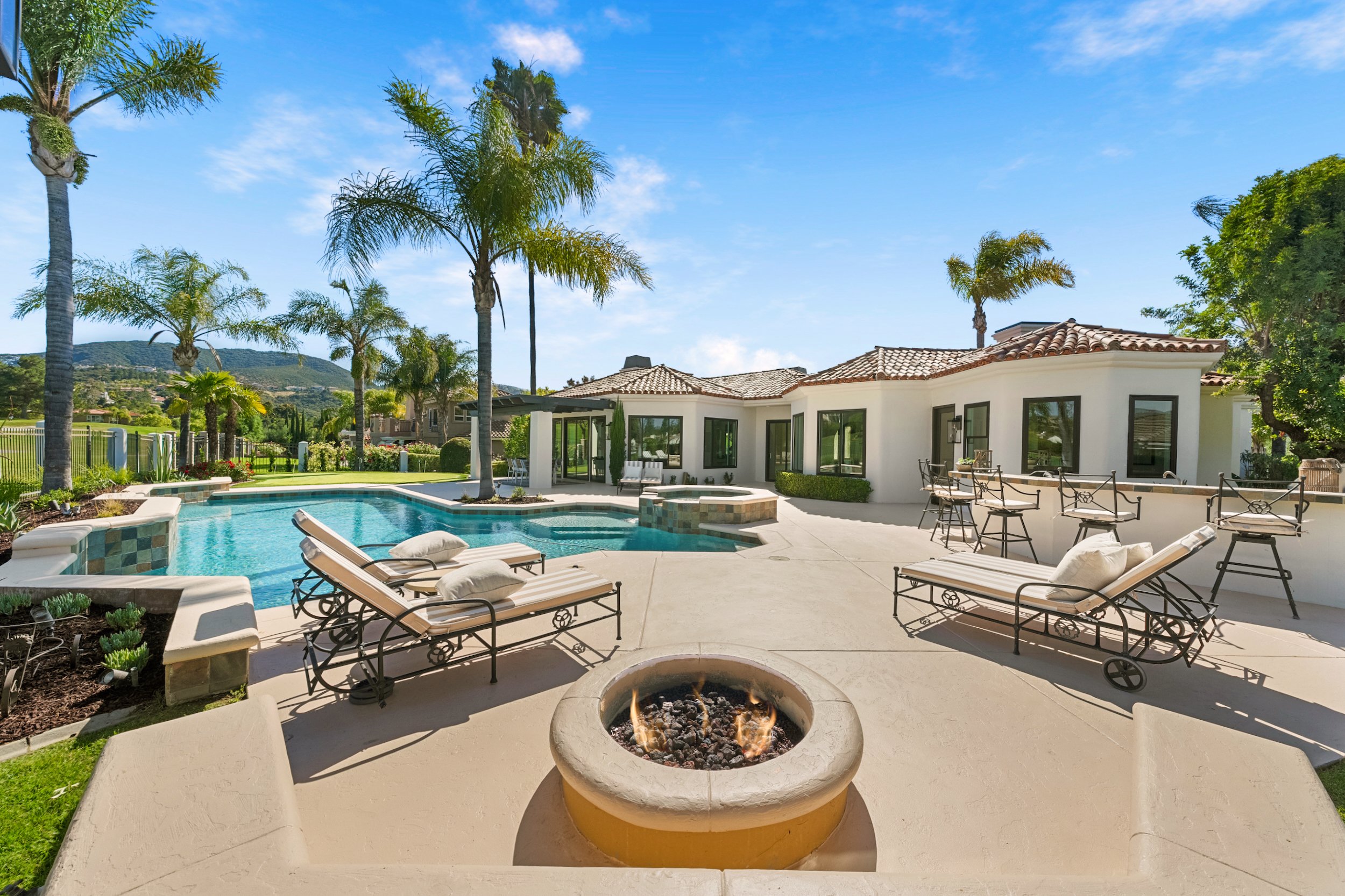 A sunny backyard with a swimming pool, lounge chairs, a fire pit, palm trees, and a modern house in the background.