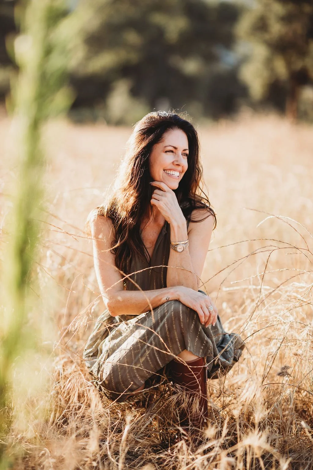 A woman sitting in a field of tall, golden grass, smiling and looking to her side, with sunlight illuminating her and a blurred background of trees.