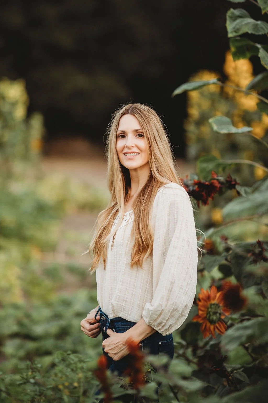 A young woman with long blonde hair and a light-colored blouse standing in a garden filled with orange and red flowers, with greenery and blurred background in the distance.