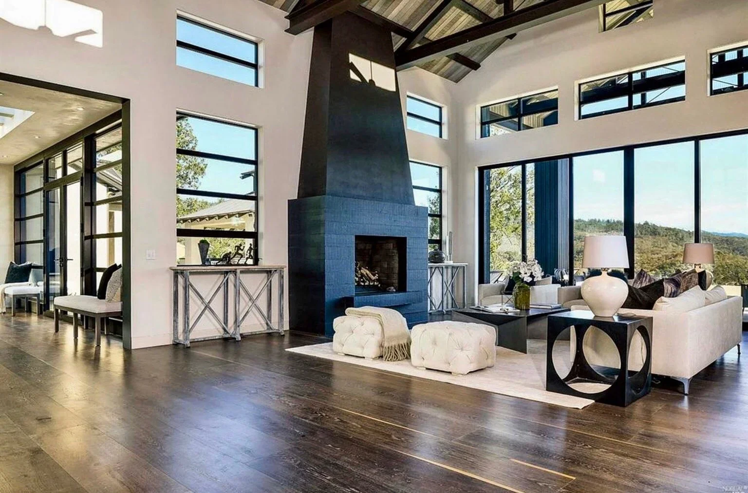 Modern living room with a fireplace, large windows, white sofas, black side tables, and wooden flooring.