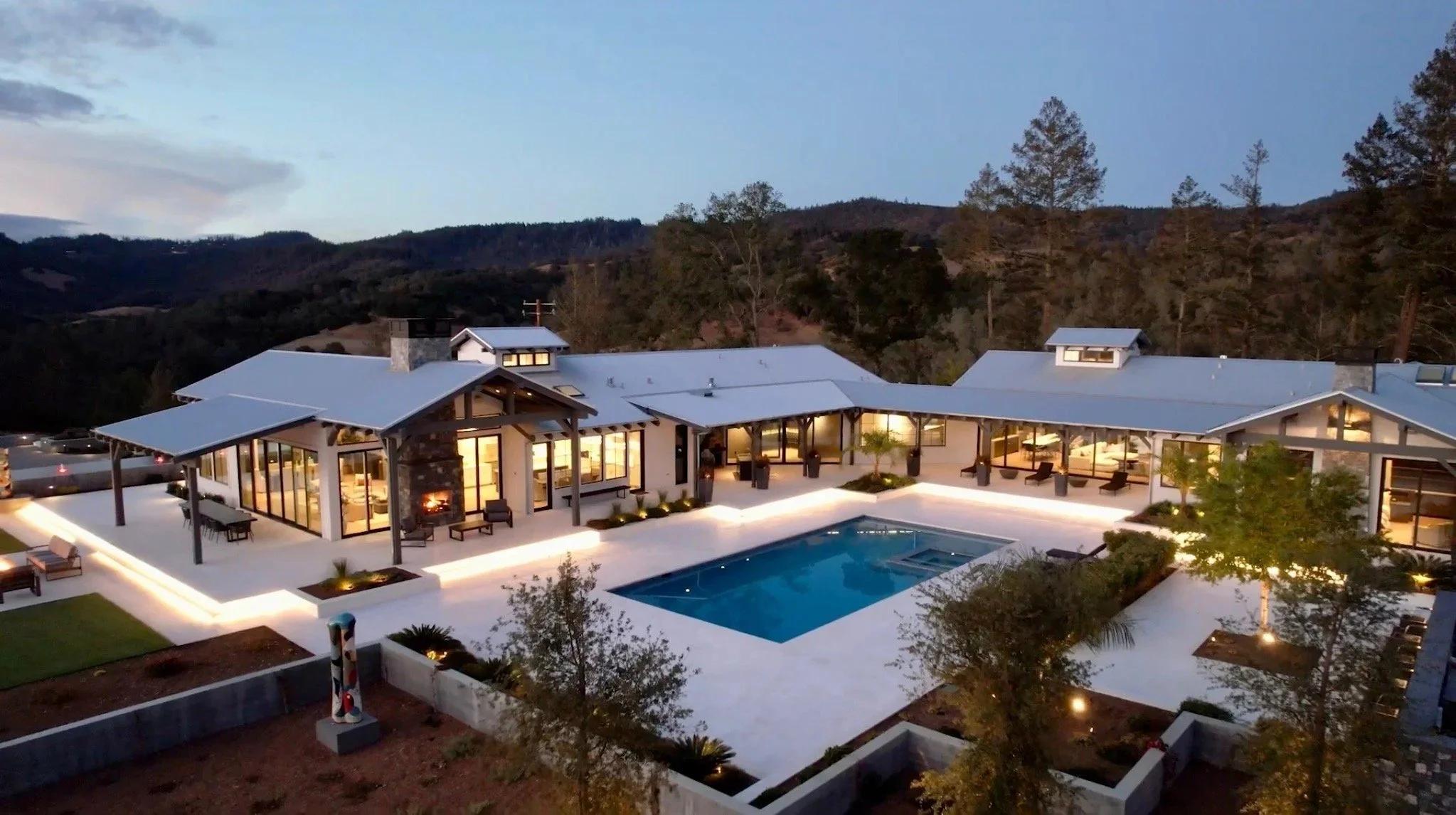 A modern house with large windows, a swimming pool, and outdoor seating, set against a backdrop of trees and hills at dusk, with interior lights visible and outdoor lighting illuminating the patio.