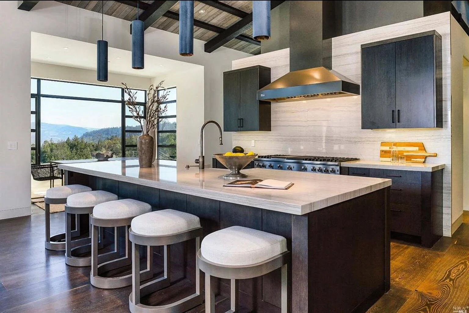 Modern kitchen with dark cabinets, marble island with seating, stainless steel appliances, large window overlooking mountains, decorative vase with branches, and open book on the island.