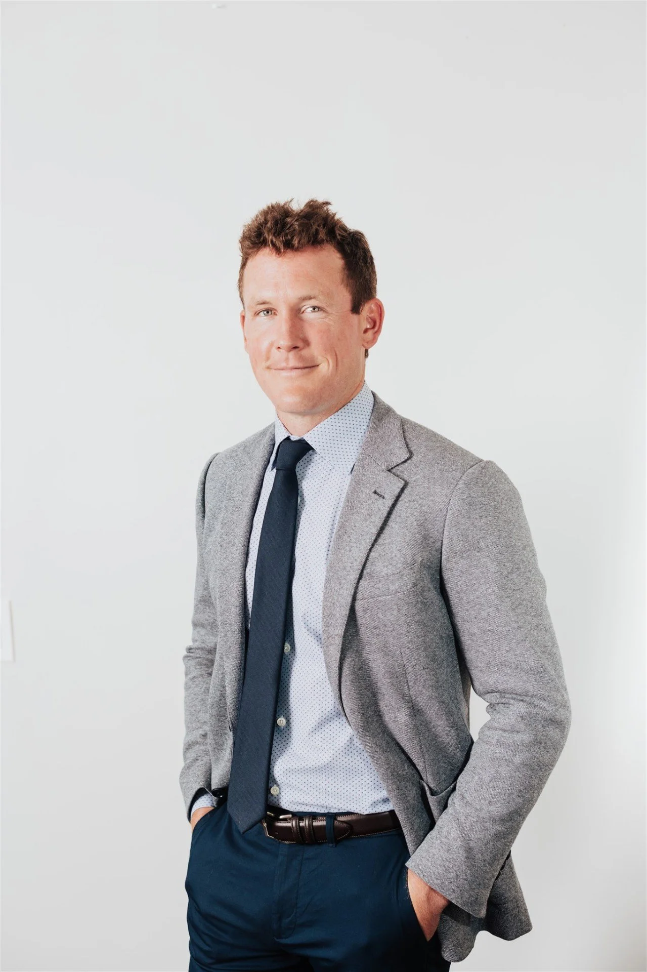 A young man in a gray blazer, light blue dress shirt, and navy tie posing with his right hand in his pocket against a plain white background.