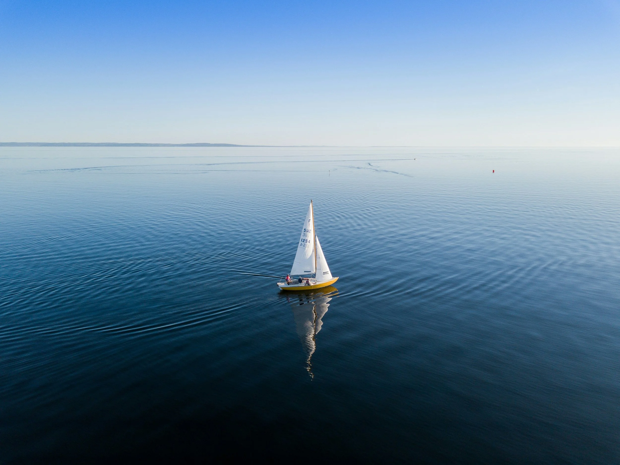A sailboat is sailing on a calm blue body of water, with a clear sky above and distant land on the horizon.