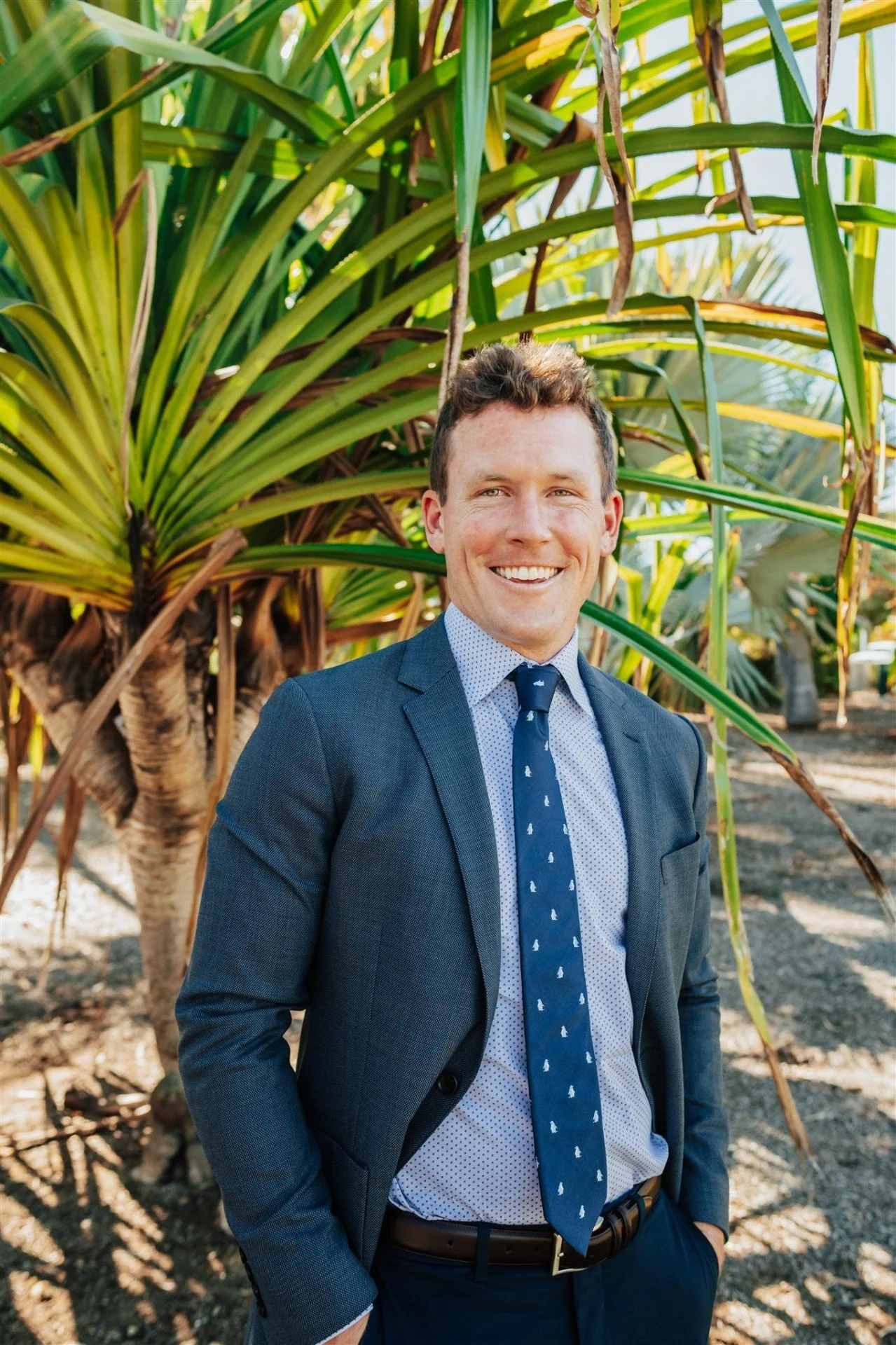 A smiling man in a business suit standing outdoors in front of large tropical plants.