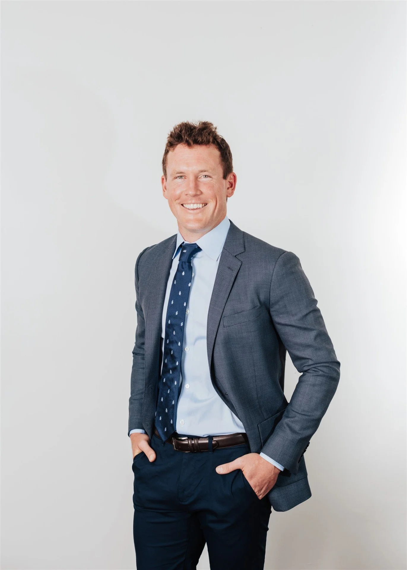 A smiling man with short, curly brown hair, dressed in a blue suit, light blue shirt, and a navy tie with white pattern, standing against a plain white background, with one hand in his pocket and the other resting on his hip.