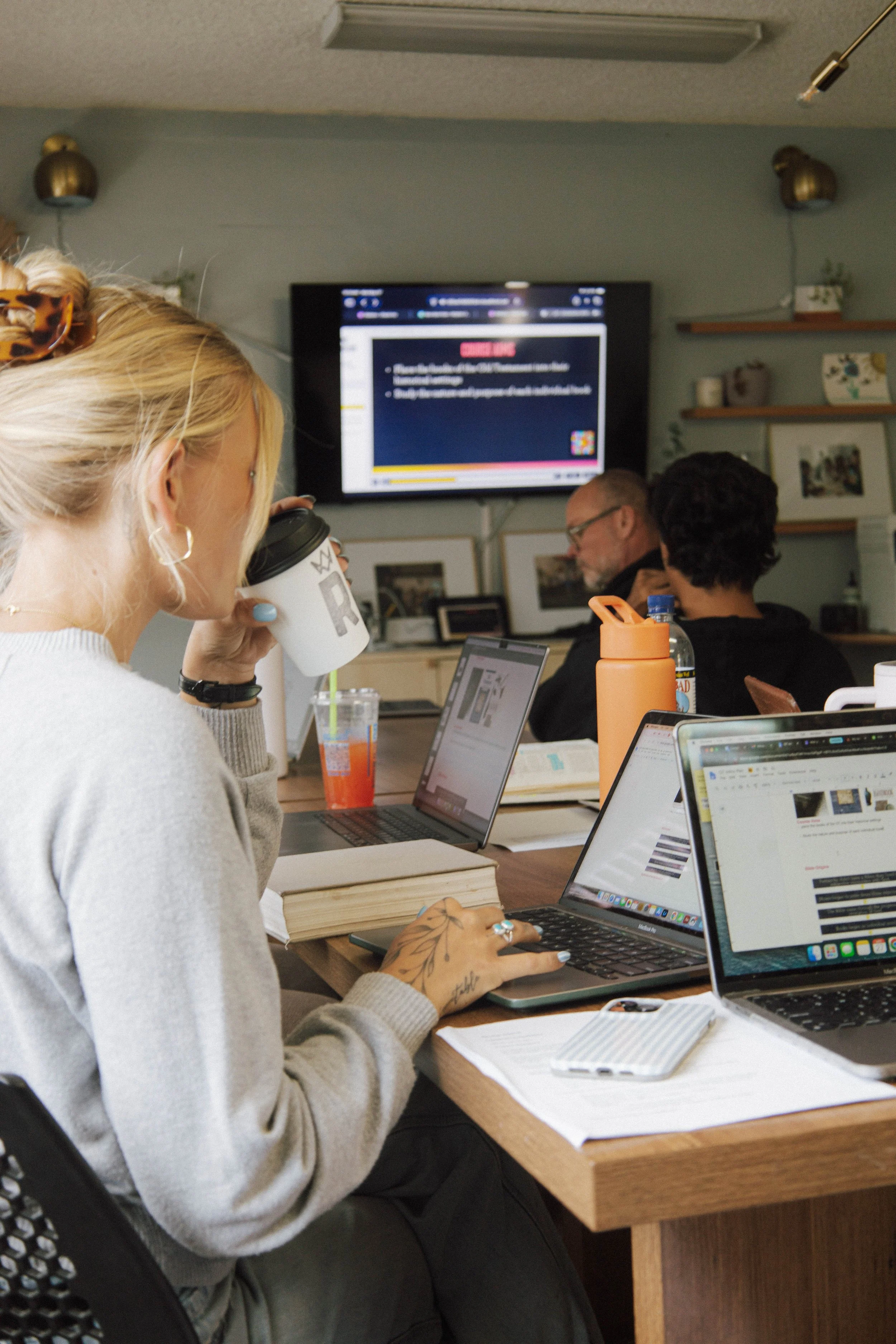 A woman with blonde hair and tattoos on her hand sitting at a table, drinking coffee, working on a laptop in a room with multiple people, computers, and a large TV screen on the wall showing a presentation.