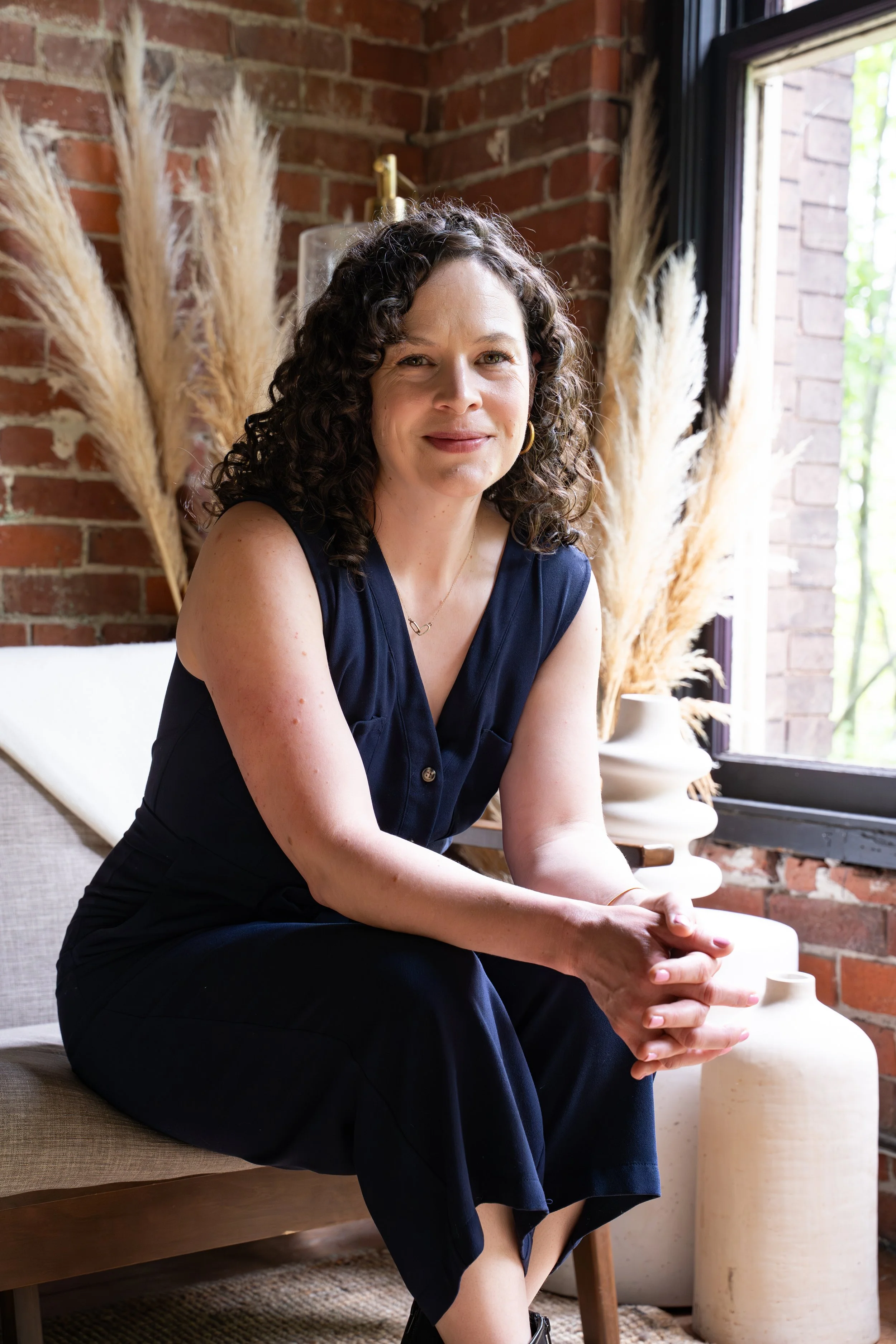 A woman with curly dark hair and light skin sitting on a beige cushioned bench near a large window with black trim, beside a brick wall with decorative pampas grass in the background, wearing a black sleeveless dress, smiling slightly, with her hands clasped on her lap.