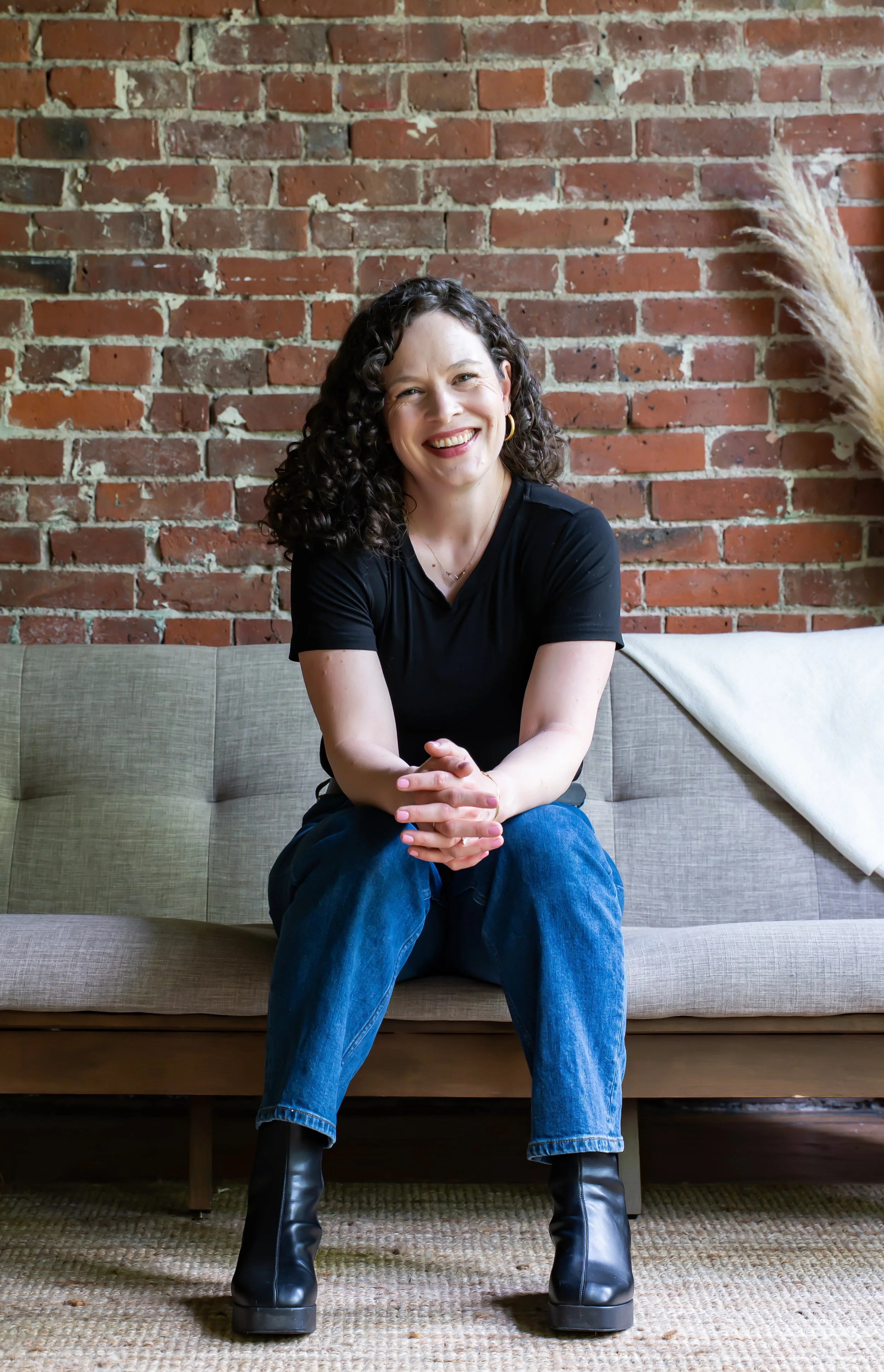 A woman with curly dark hair, wearing a black shirt, blue jeans, and black boots, sitting on a beige couch in front of a brick wall, smiling at the camera.