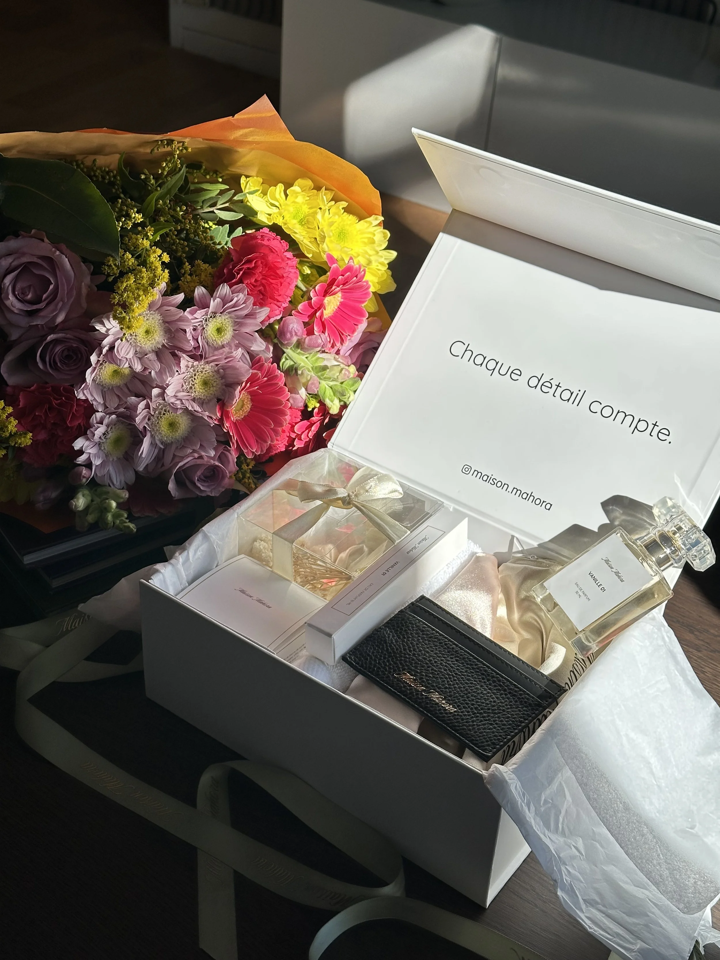 Un bouquet de fleurs colorées posé près d'une boîte blanche contenant des produits de parfum, comme des flacons et une carte de vœux, sur une surface en bois.