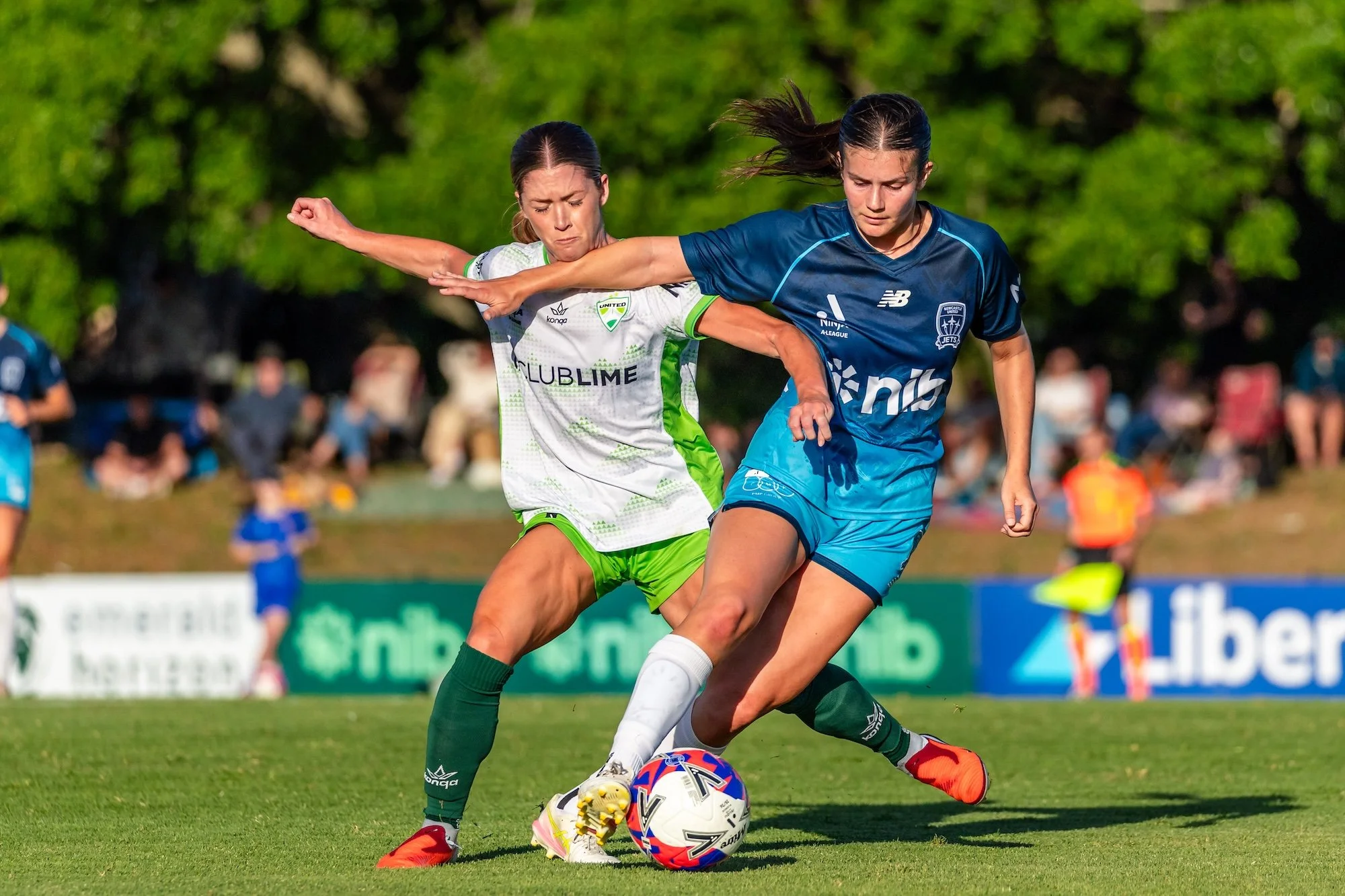 Two women’s soccer players competing for the ball on a grassy field, one in a blue Newcastle Jets uniform and the other in a white and green uniform.