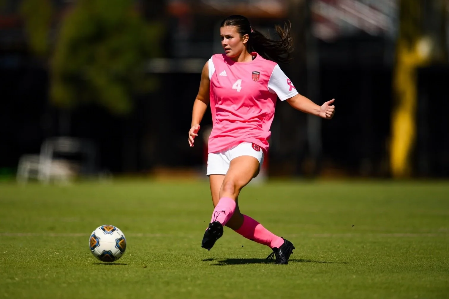 Female soccer player wearing a pink No. 4 jersey running toward a soccer ball on a field