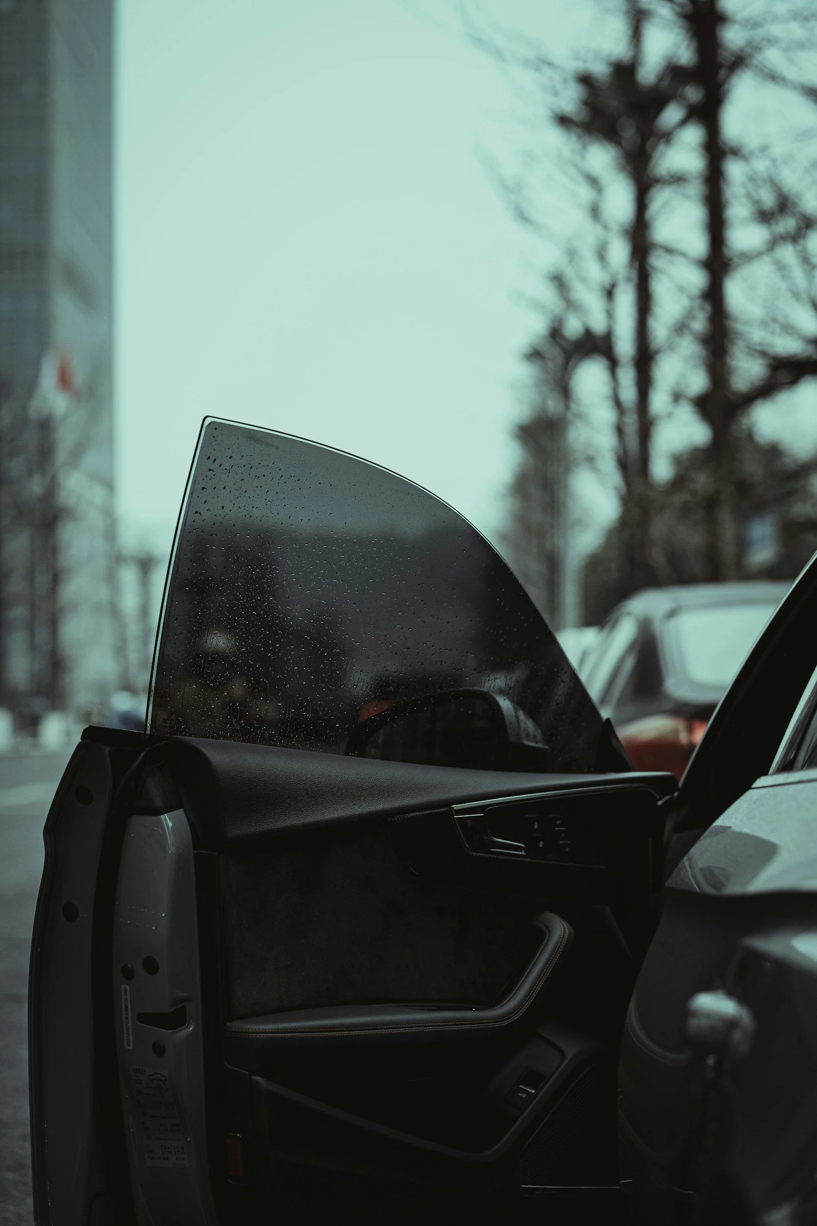 Inside perspective of a car showing the open front door with the dashboard and part of the driver's side door visible, and a slightly fogged S-curve windshield with raindrops, with trees and vehicles outside in a parking lot.