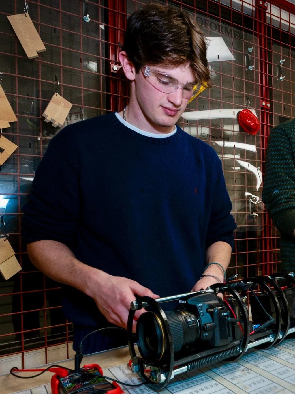 Ethan Arterburn in glasses working with a Terrapin 1 aquatic camera trap in a in a workshop or lab, with a red grid wall and various small items and notes behind him.