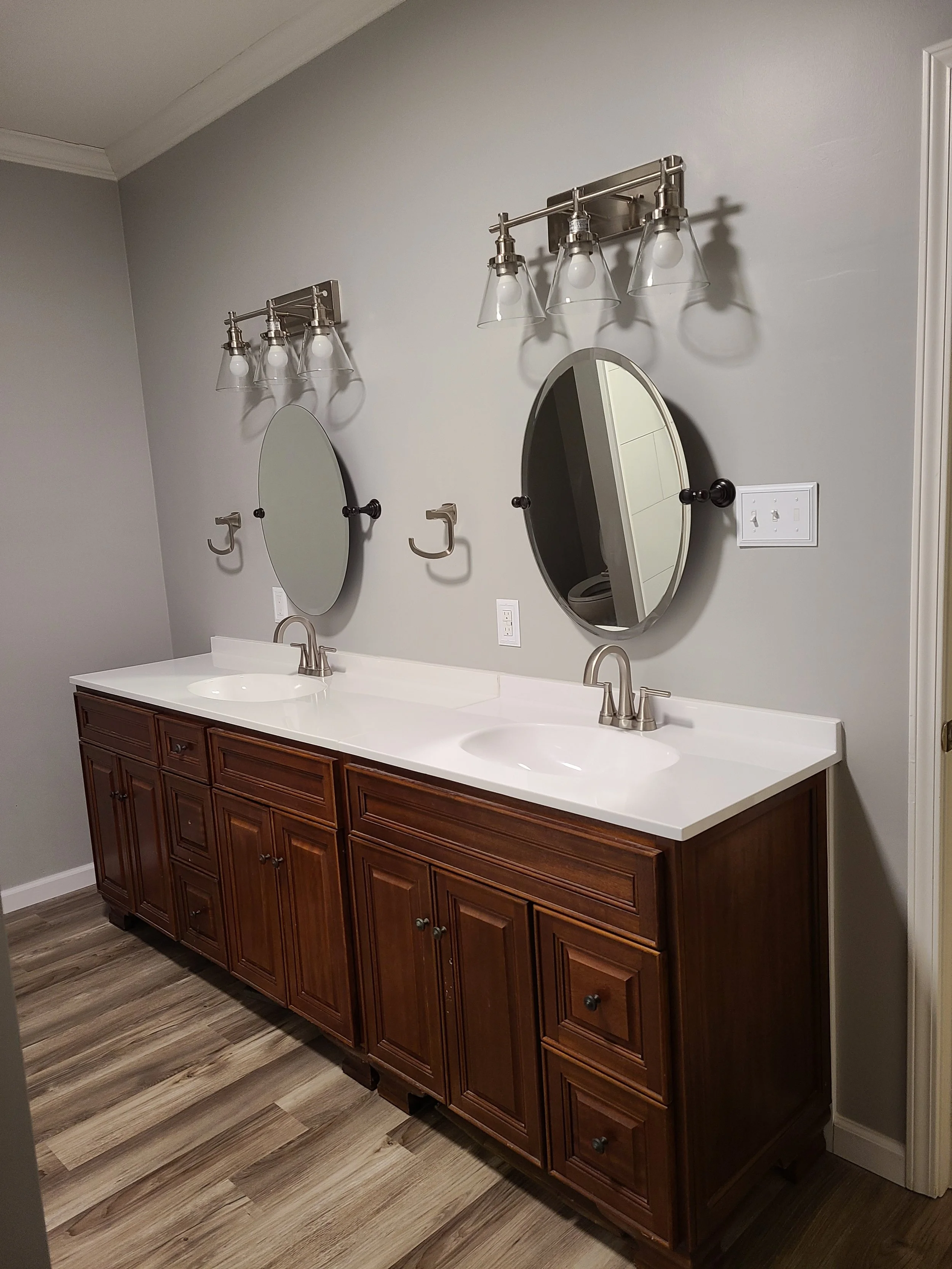 Bathroom vanity with double sinks, wooden cabinets, oval mirrors, wall-mounted light fixtures, and a light switch.