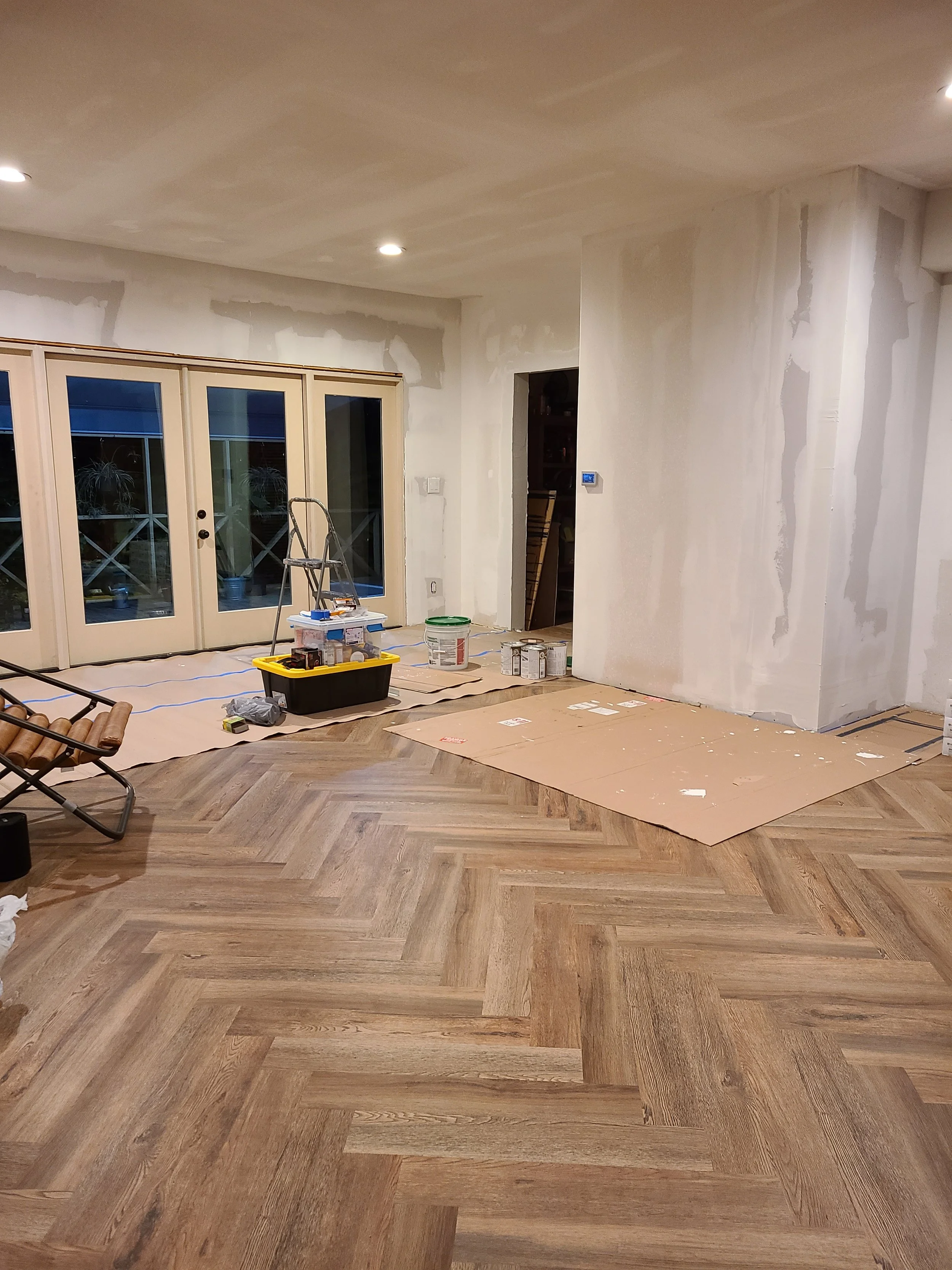Interior of a house under renovation showing a newly installed herringbone wood floor, partially painted walls, tools, and materials on the floor.