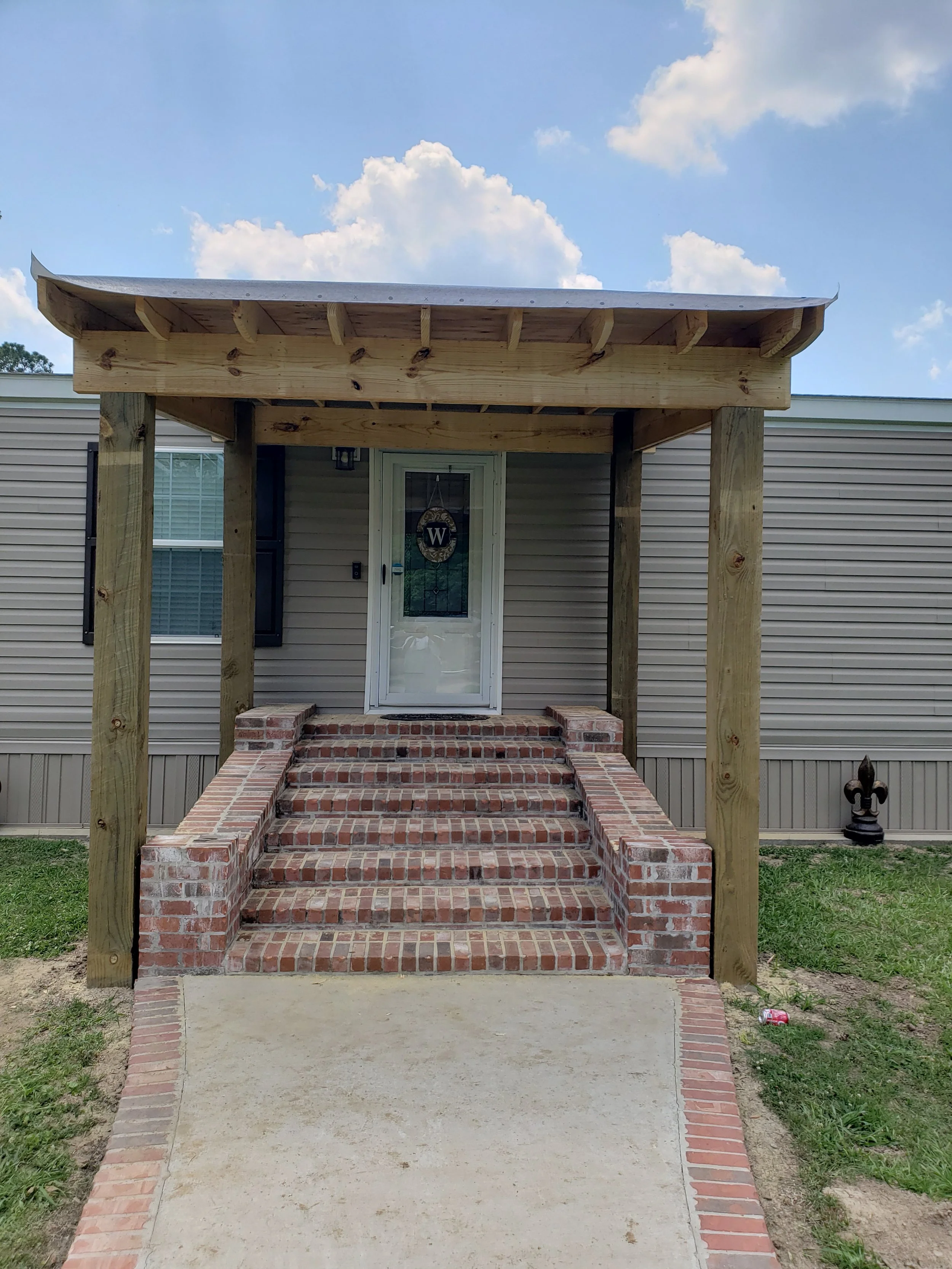 A house entrance with brick stairs, a concrete walkway, a white door with a decorative wreath, a covered porch area with wooden supports and a metal roof, and a beige vinyl siding exterior.