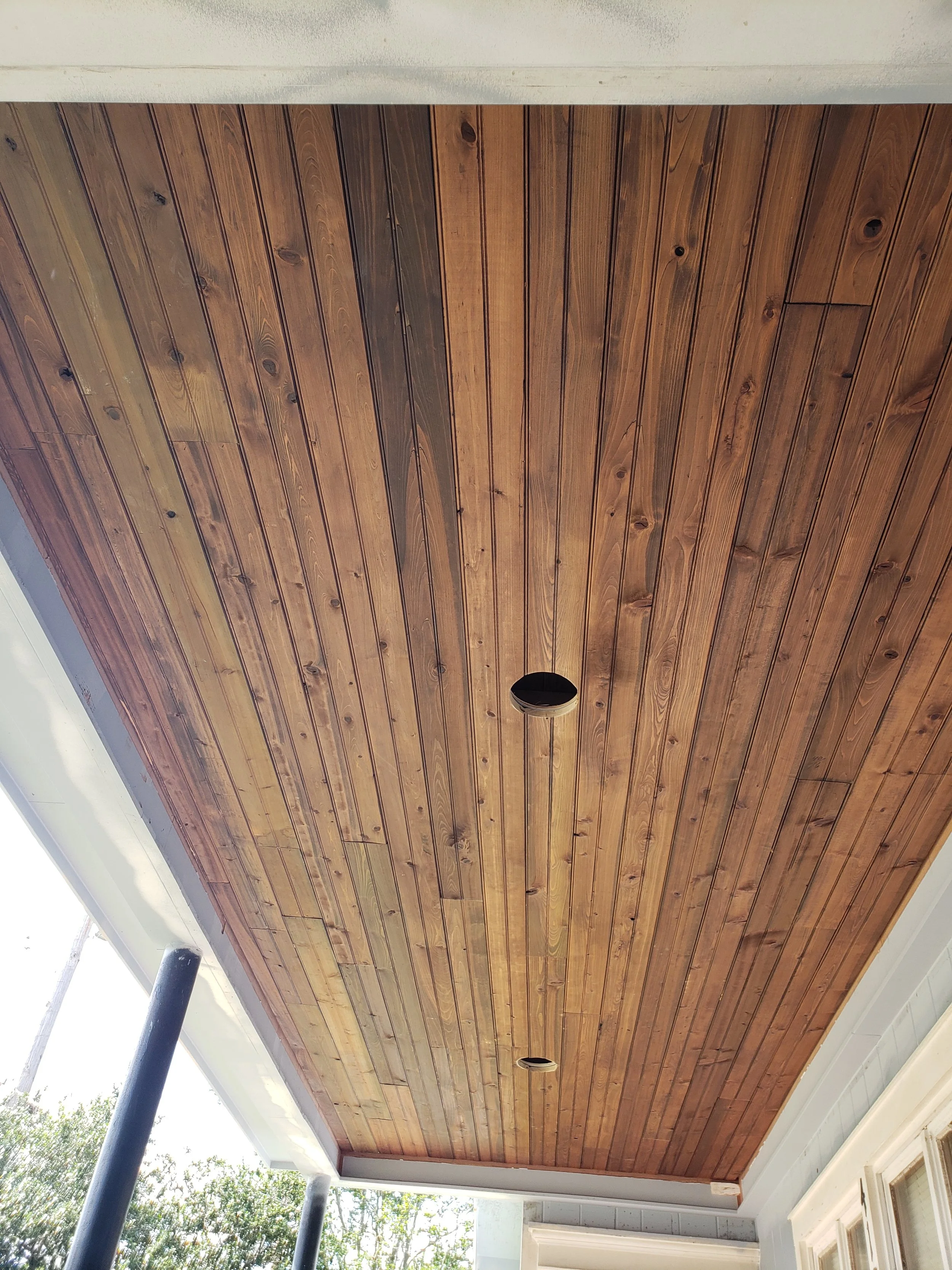 Wooden tongue and groove plank ceiling with holes for recessed lighting, supported by black metal pipes, beside white walls and windows.