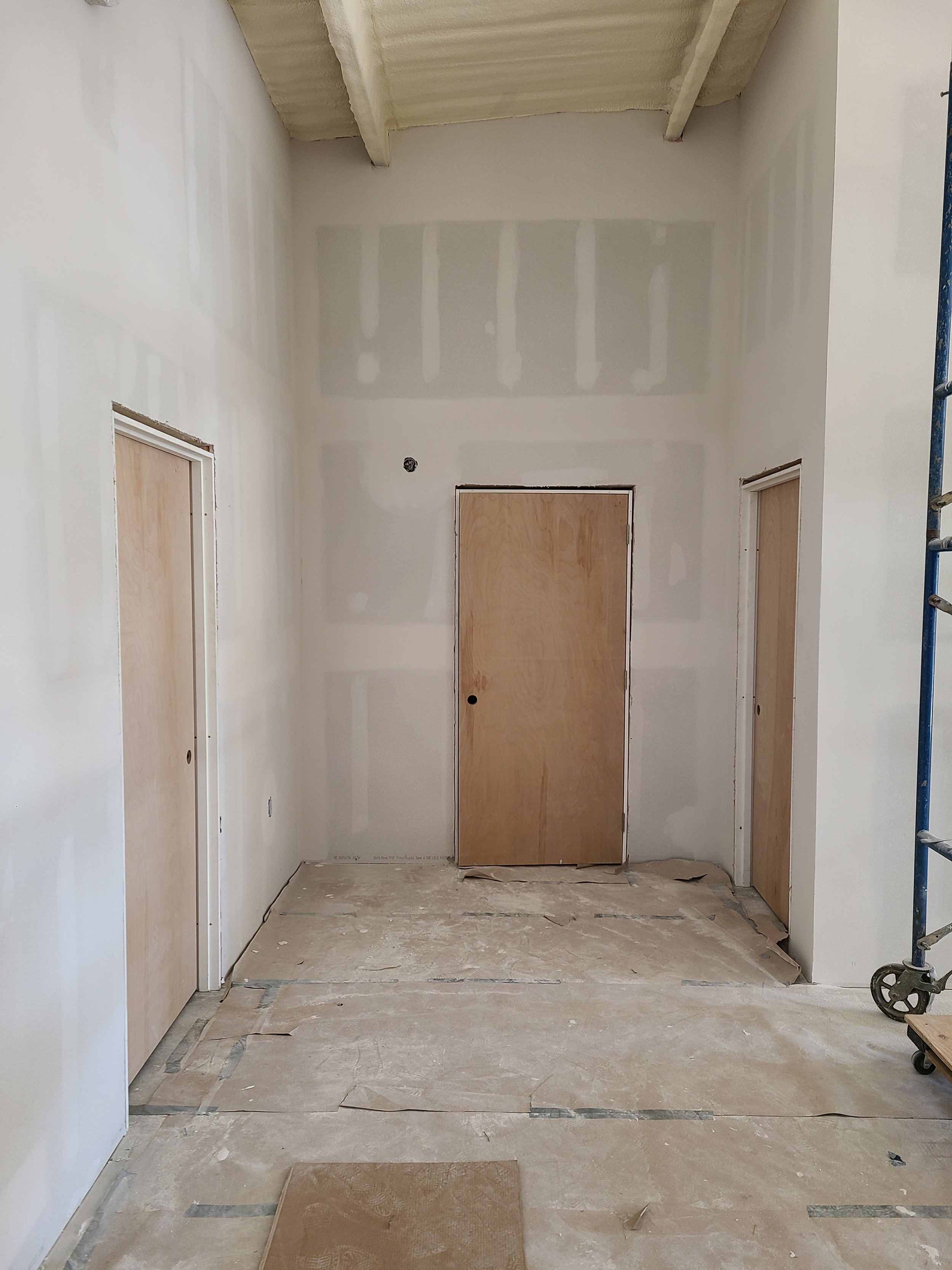 Interior view of a hallway under construction with three unfinished doorways, unpainted drywall, and protective covering on the floor and doorway frames.