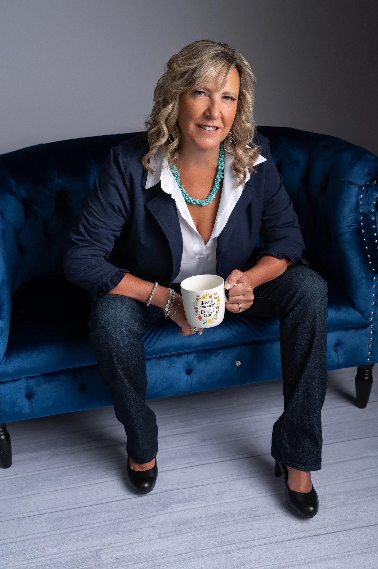 A woman with blonde, curly hair sitting on a blue velvet sofa, holding a white mug with floral design and inspirational quotes, wearing a navy blazer, white shirt, black jeans, black heels, turquoise necklace, and silver jewelry, smiling and looking at the camera.