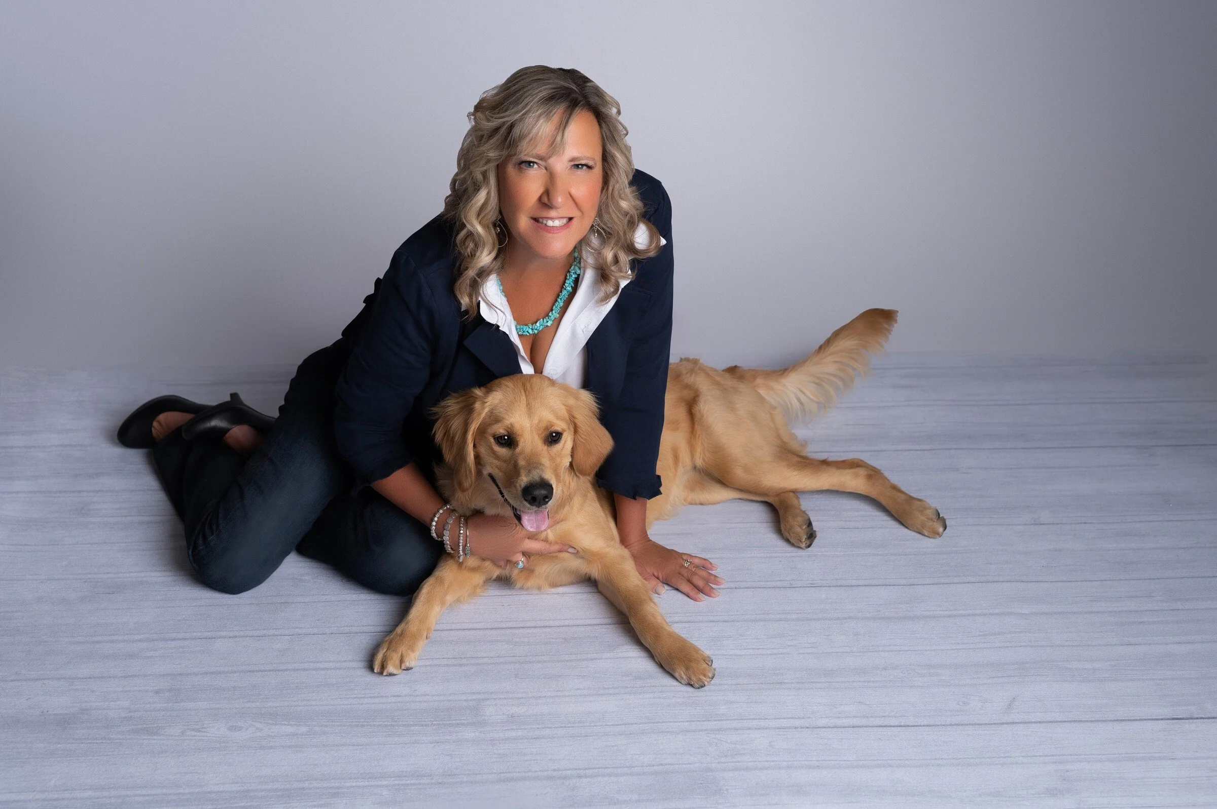 A woman with blonde curly hair squatting on the floor next to a golden retriever lying down, both looking at the camera against a plain light gray background.