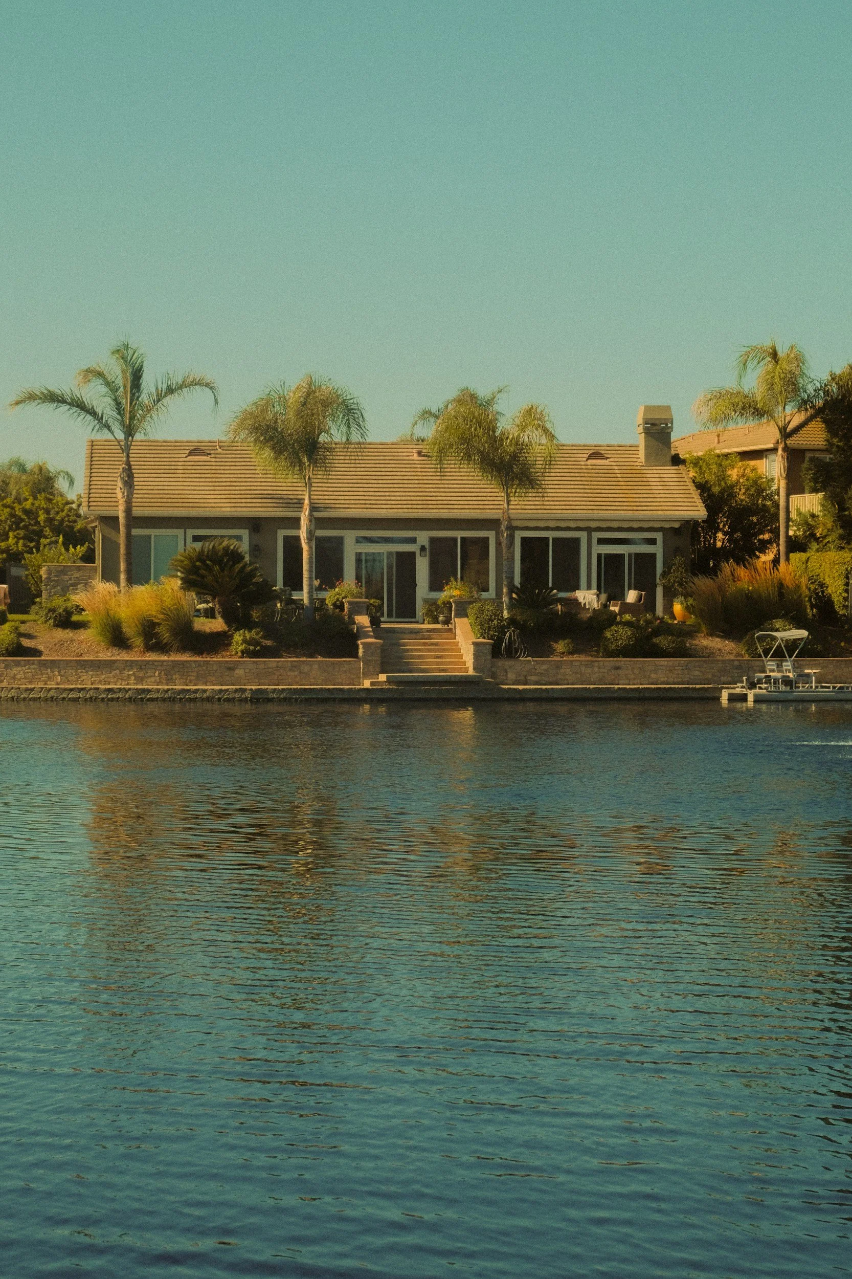 DOMUS Home Watch Services - A house located by a body of water with a sandy shoreline, palm trees in front, and a clear sky overhead.