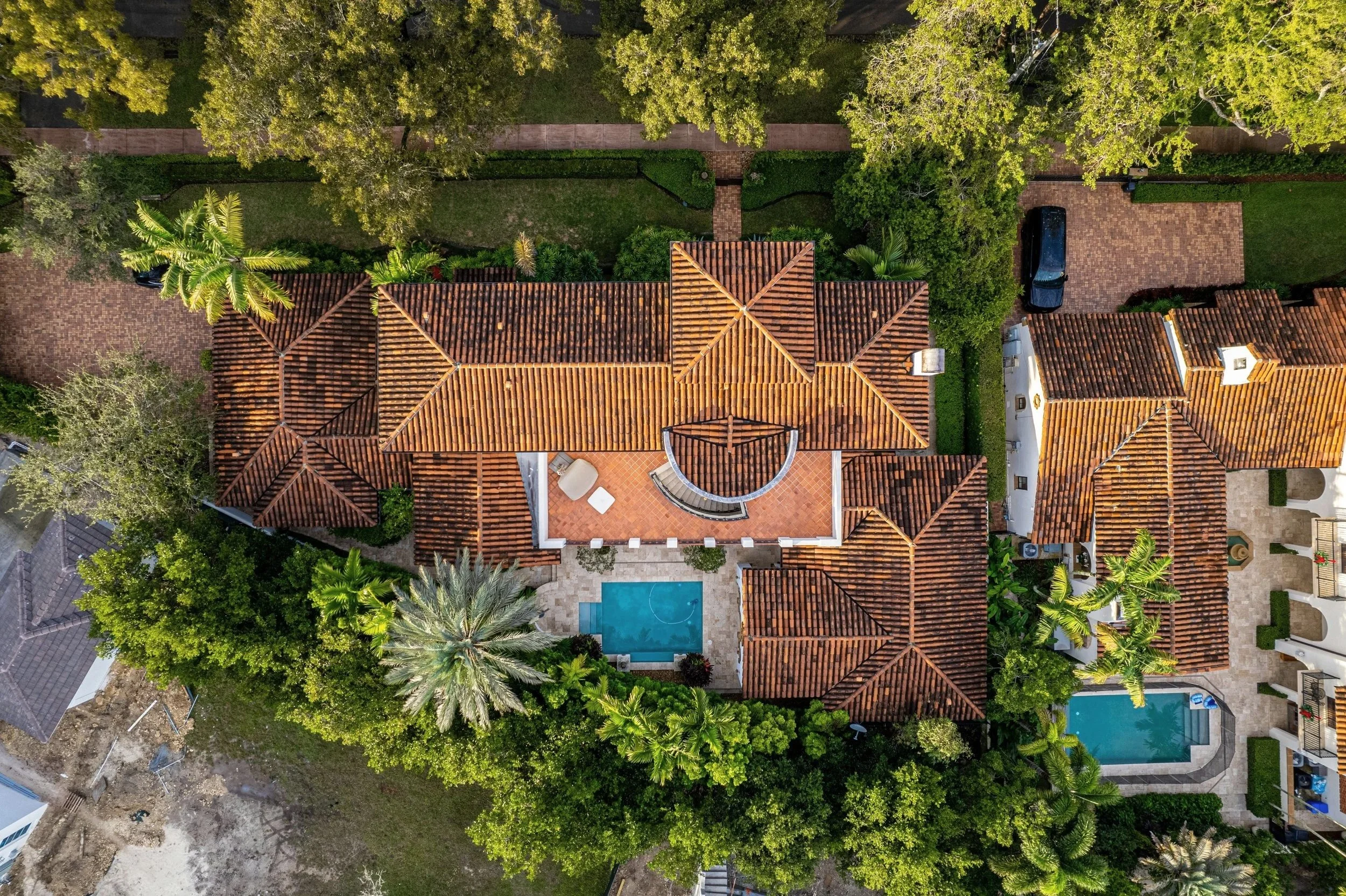 DOMUS Home Watch Services - Aerial view of a residential area with multiple houses featuring red-tiled roofs, green trees, a swimming pool, and several parked cars.
