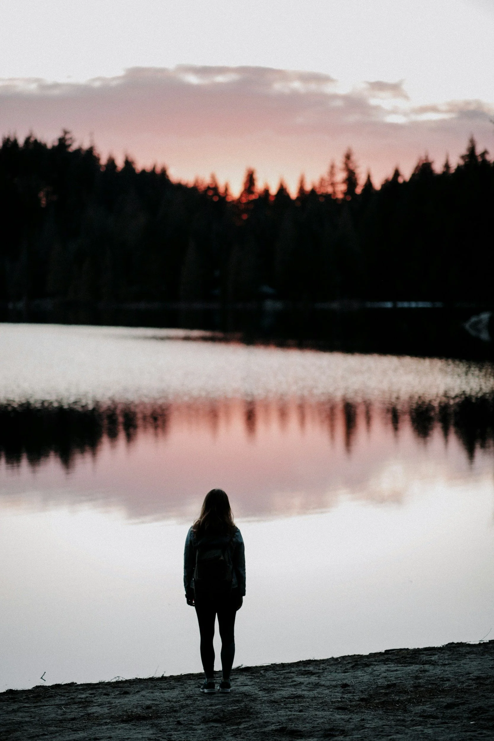 A person standing on the rocky shoreline of a lake or river at dusk, gazing at a calm body of water with a forested tree line and cloudy sky reflected in the water.