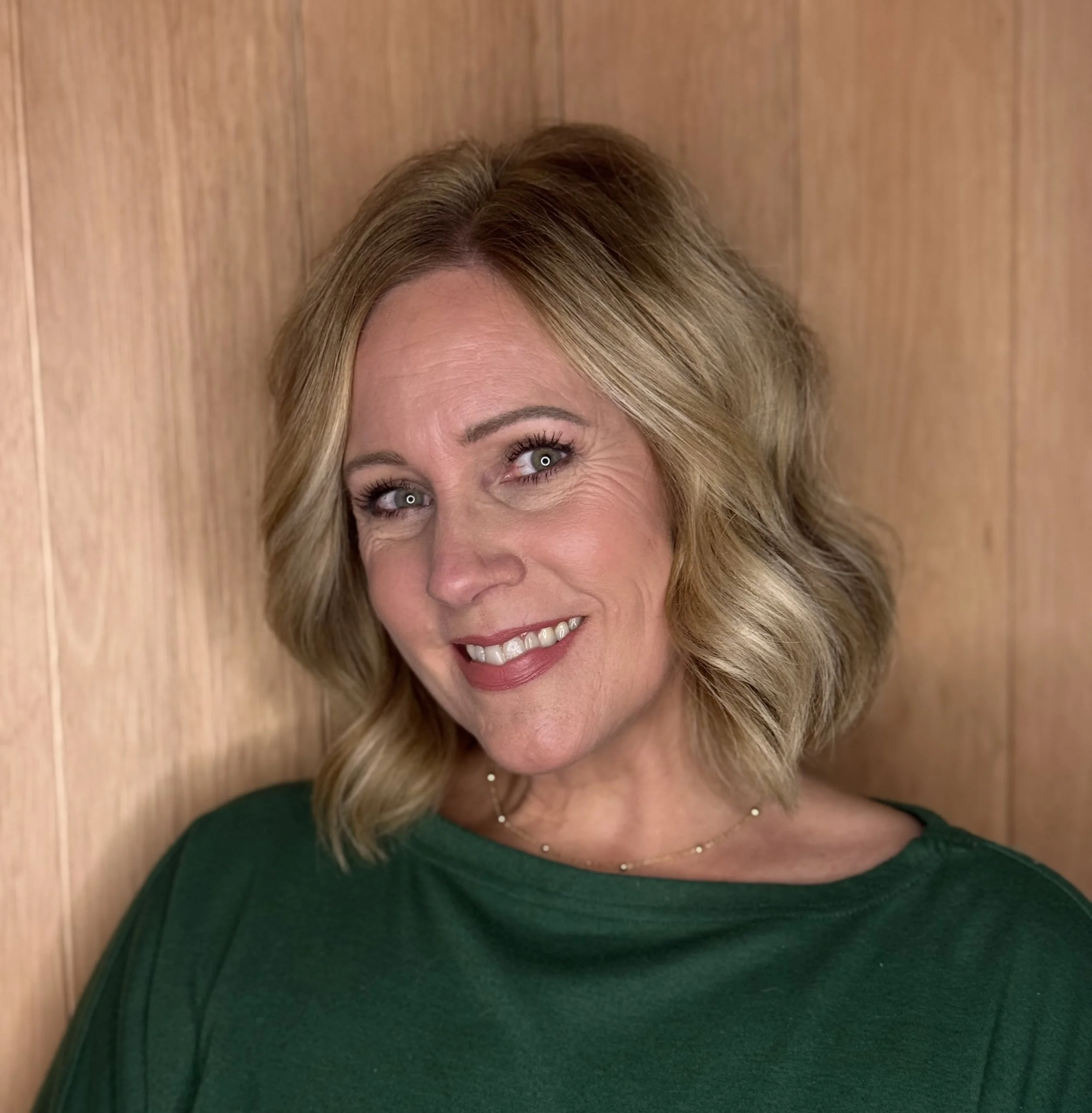 Close-up of a smiling woman with blonde hair, wearing a green top, against a wooden background.
