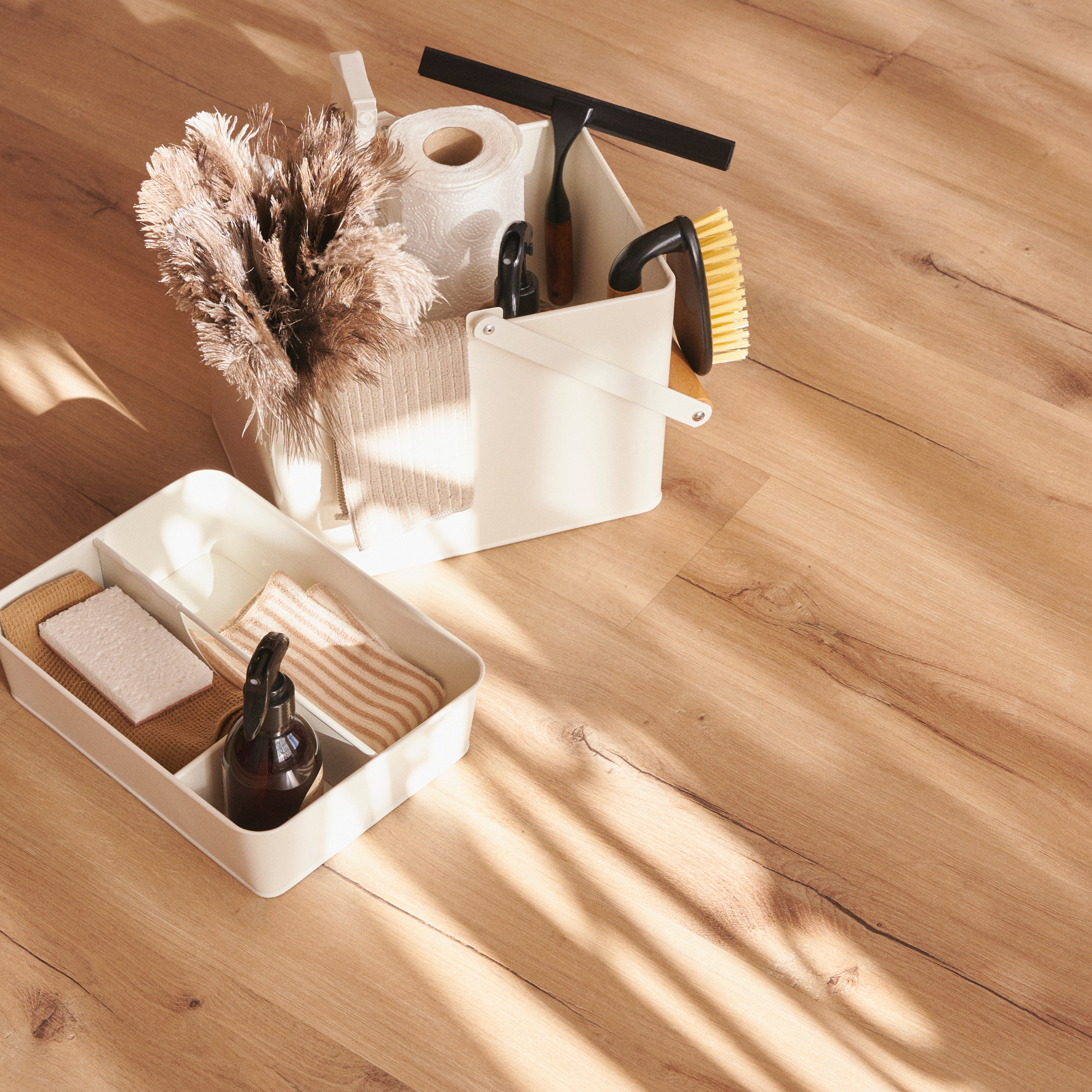 A white tank-style storage caddy on a wooden floor, holding dried beige and brown pampas grass, a roll of toilet paper, and a pair of hedge trimmers.