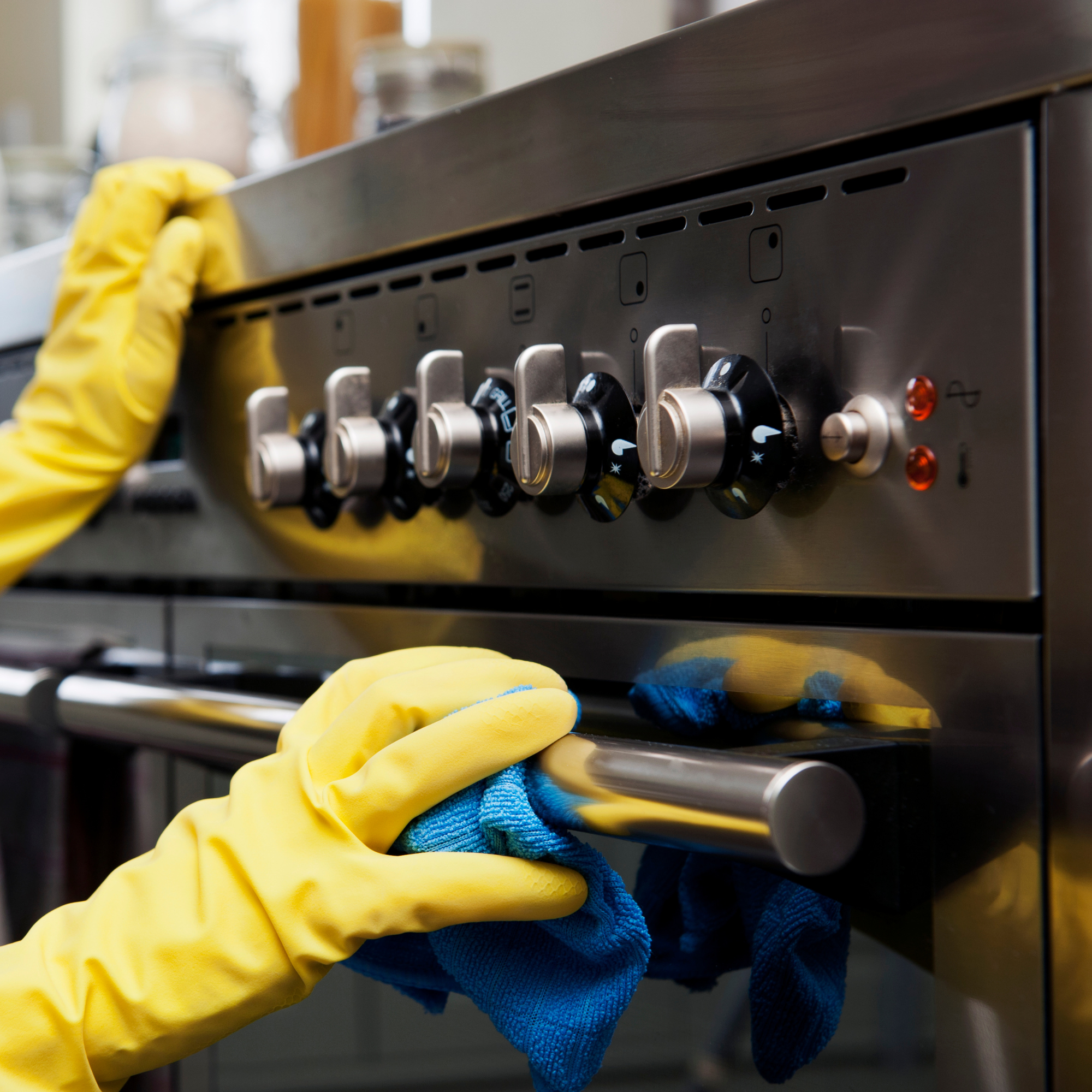 A person wearing yellow gloves is cleaning or wiping a stainless steel stove or oven surface with a blue cleaning cloth.