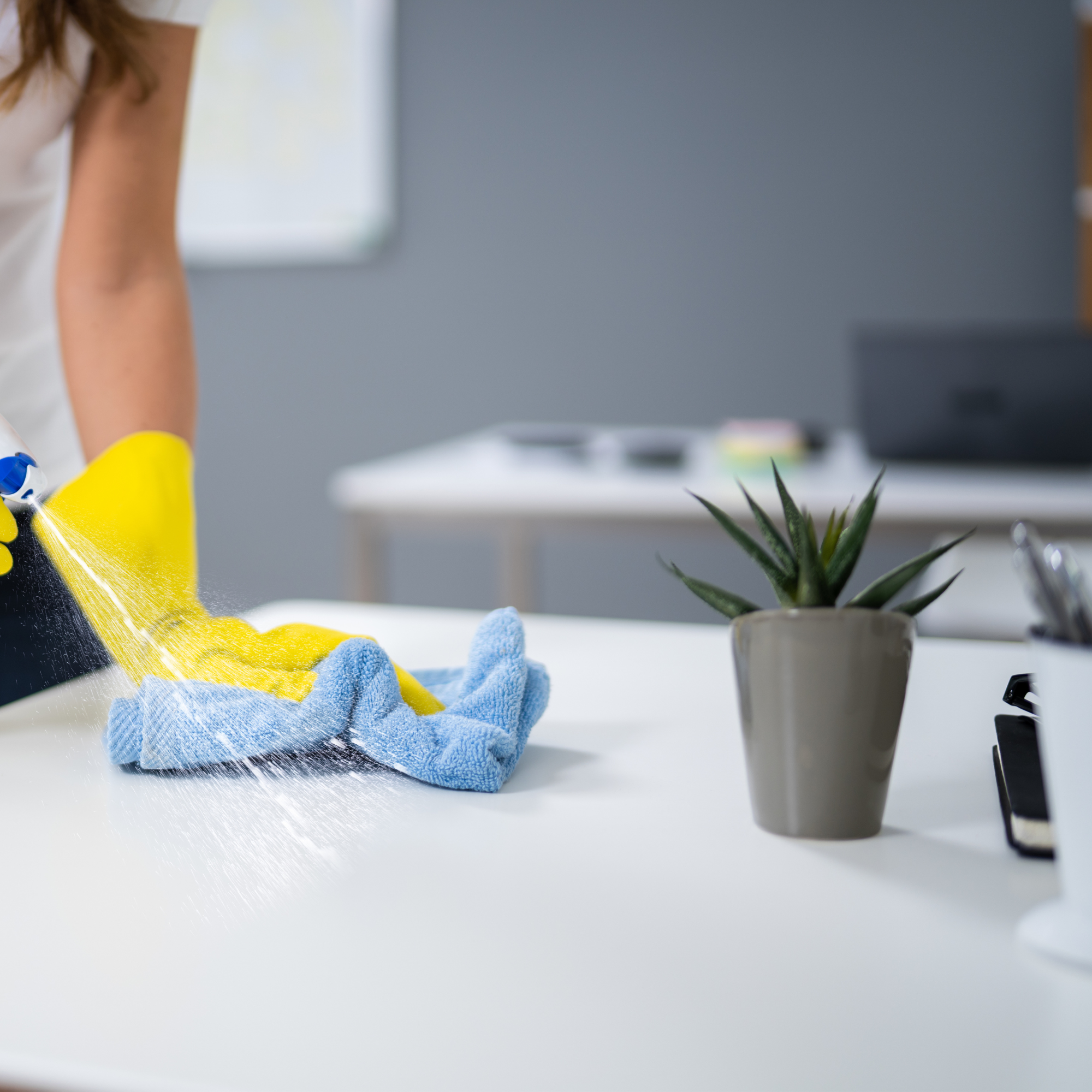 Person wearing yellow gloves cleaning a glass surface with a blue and yellow cloth, with a potted plant on the table and a blurred computer monitor in the background.
