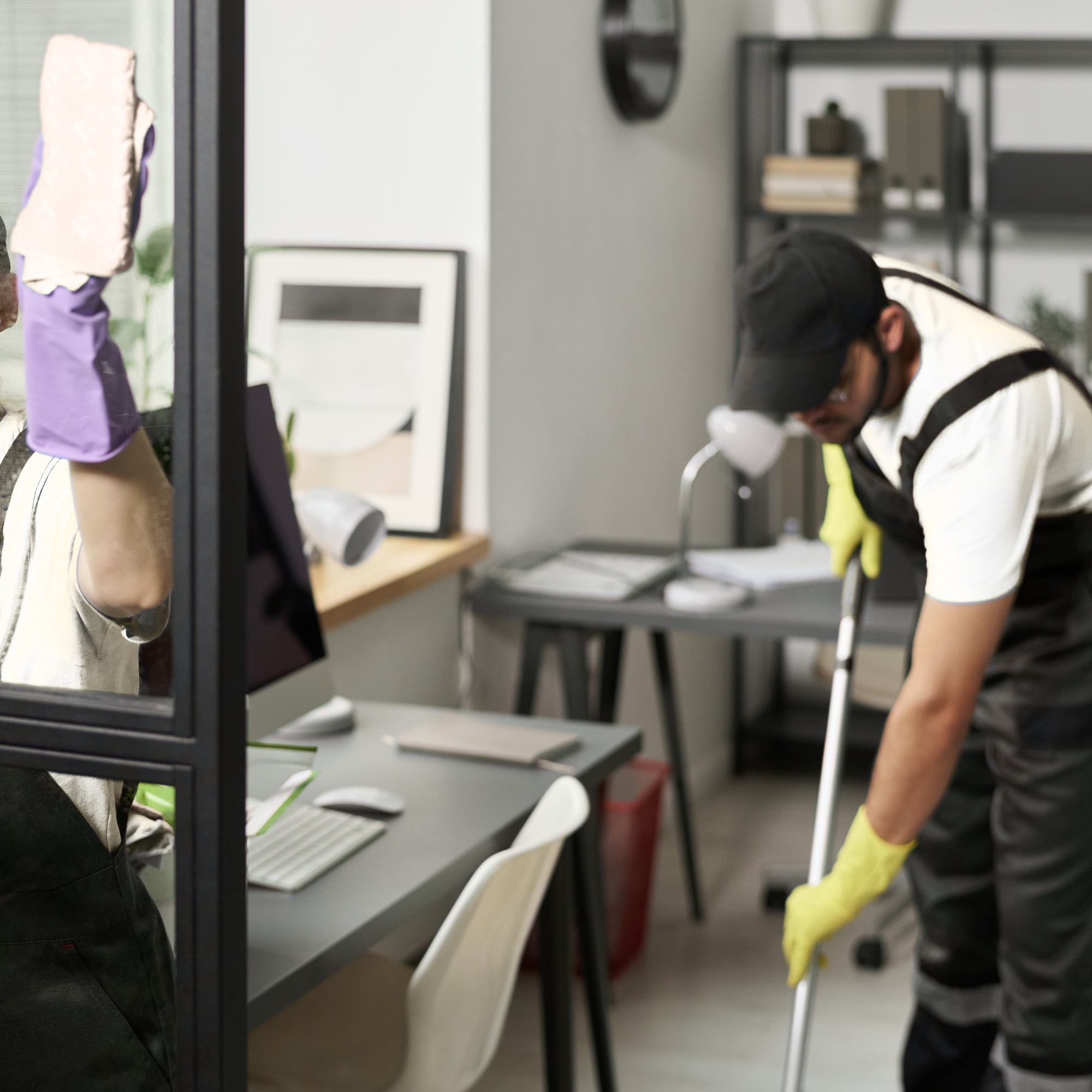 People in a modern office or co-working space, with some wearing safety vests and caps, working or discussing, with shelves and framed pictures in the background.