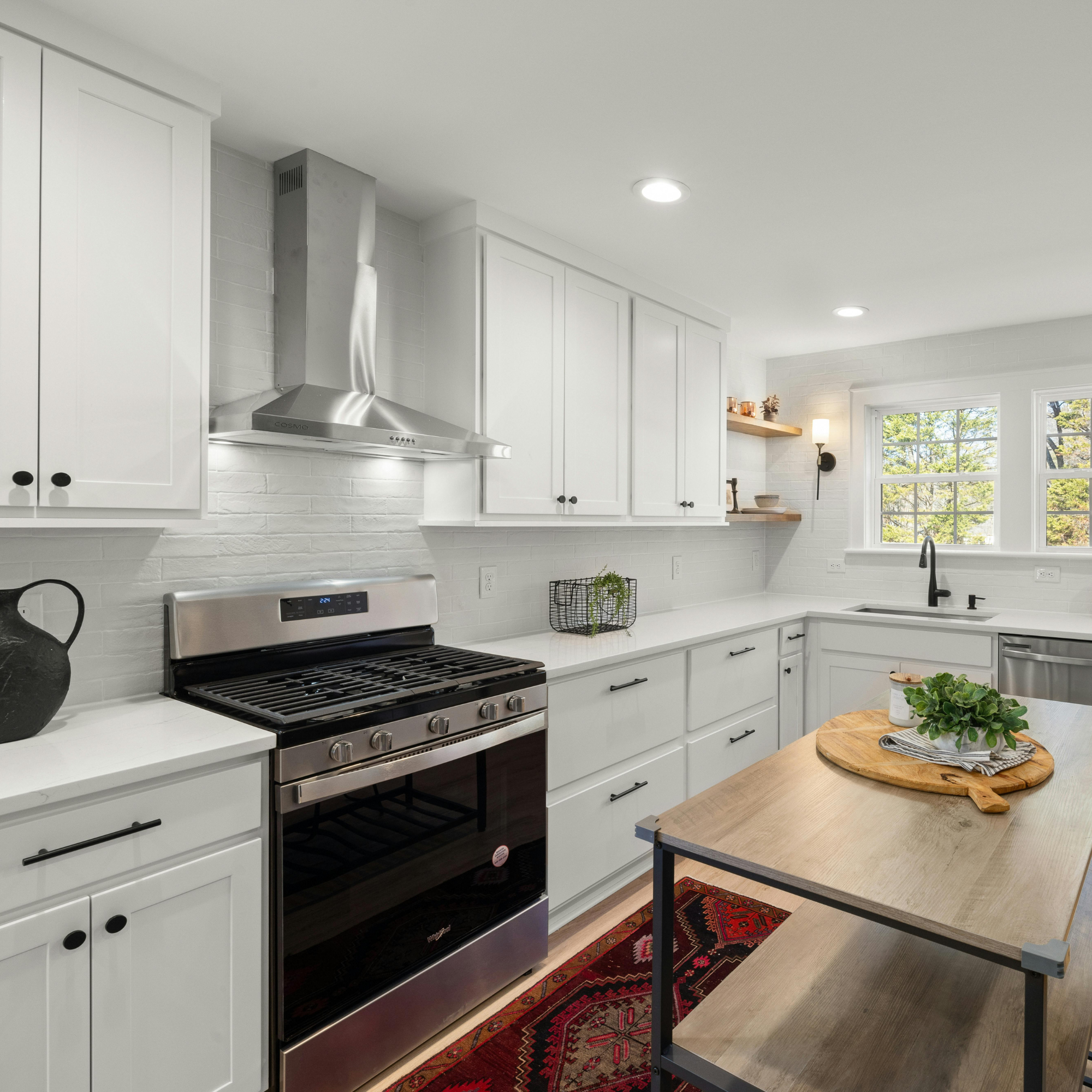Modern kitchen with white cabinets, stainless steel oven and stove, black handles, a red patterned rug, a wooden table with a potted plant, large window with green trees outside, and ceiling lights.