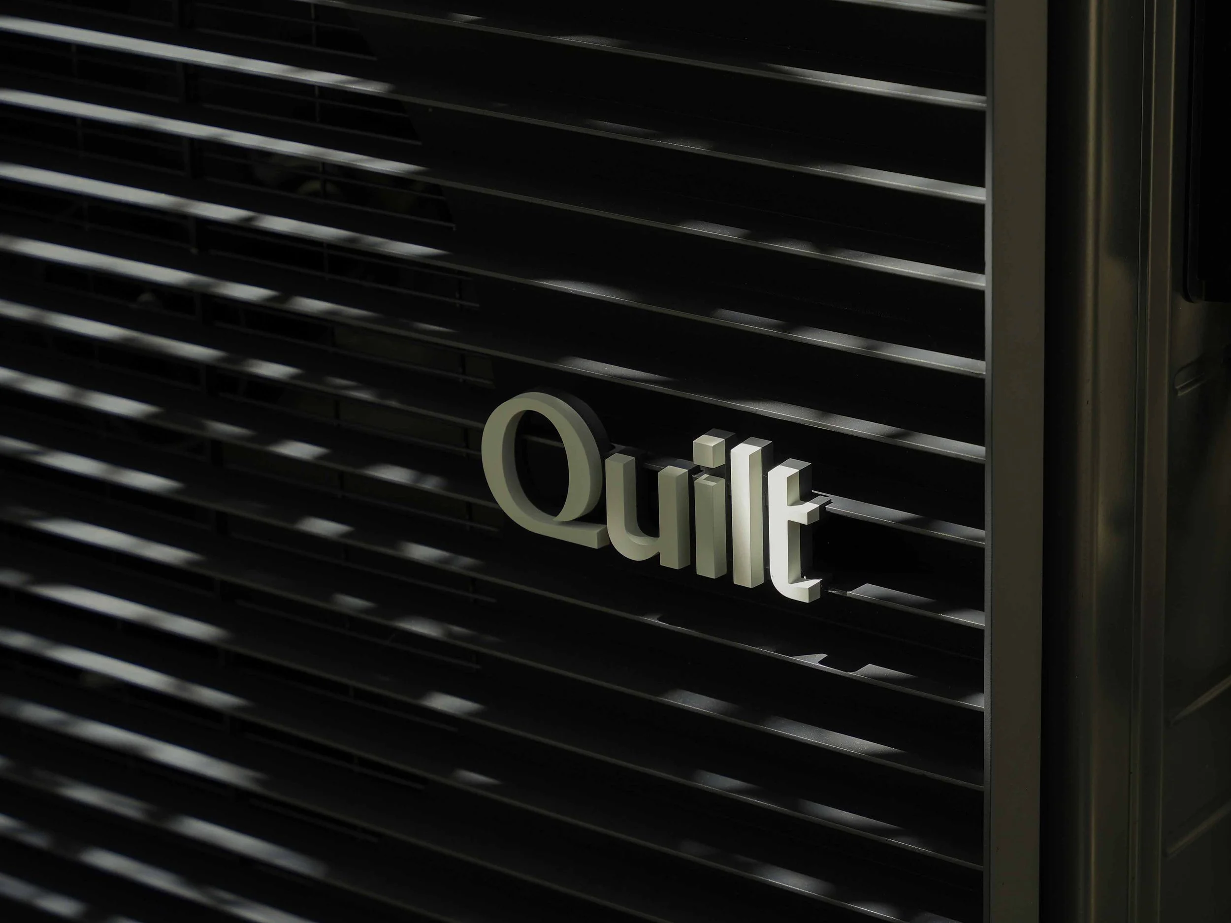 Close-up of the premium silver Quilt logo badge on the front grille of an outdoor heat pump system.