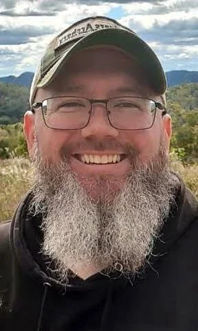 A man with glasses and a beard, smiling outdoors, wearing a baseball cap and a hoodie, with mountains and cloudy sky in the background.