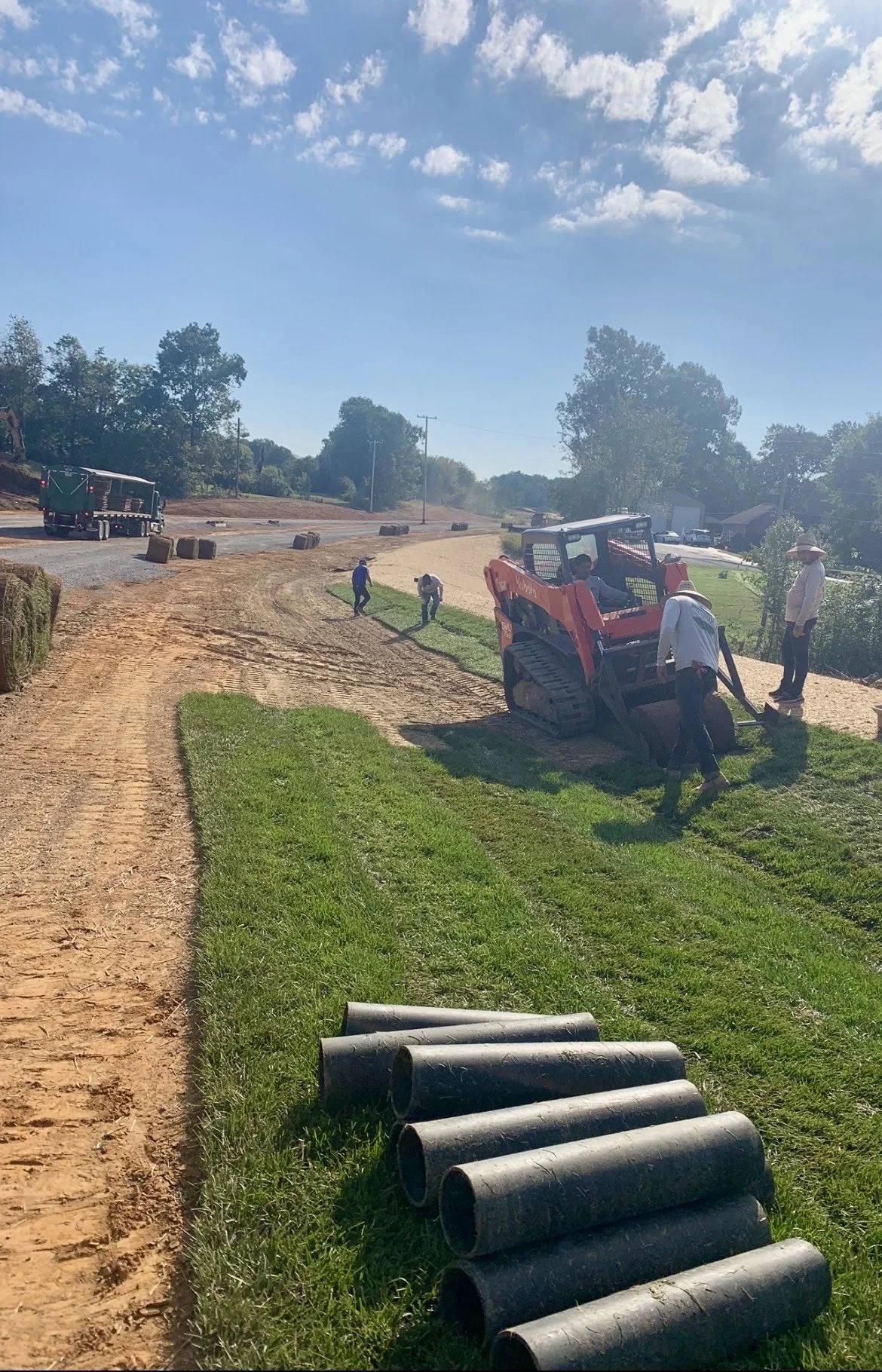 Construction workers laying down black pipes on a grassy area while operating a skid steer loader, with a dirt path and trees in the background under a partly cloudy sky.