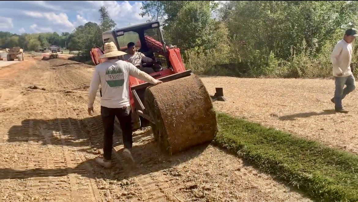 People leveling and spreading soil on a patch of grass along a dirt road with a small tractor and trees in the background.