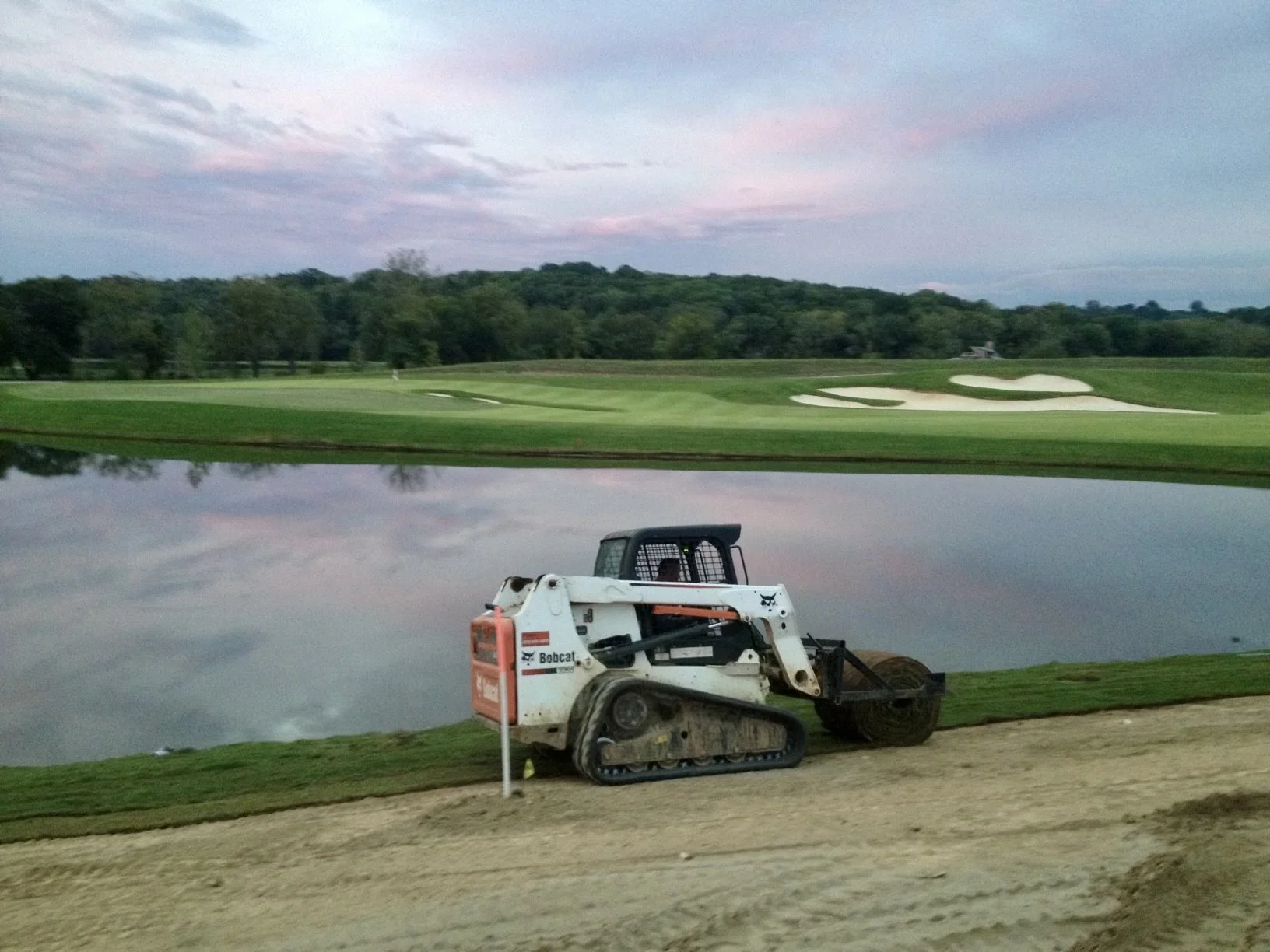 A construction vehicle near a pond at a golf course, with well-maintained green grass and sand bunkers in the distance, under a partly cloudy sky.