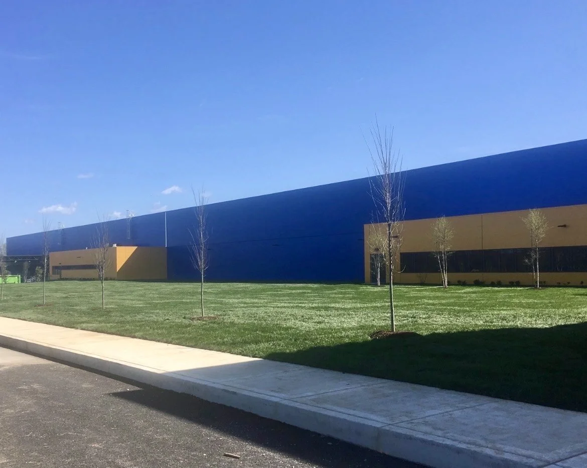 A large industrial or warehouse building with a blue and tan exterior, surrounded by a green lawn with small, leafless trees, under a clear blue sky.