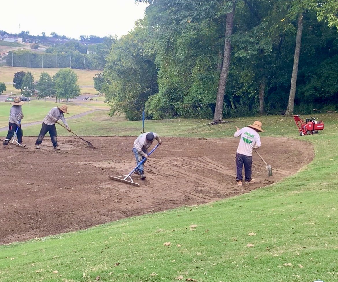 Four men are working on planting or leveling soil in a park or golf course with grass and trees in the background.