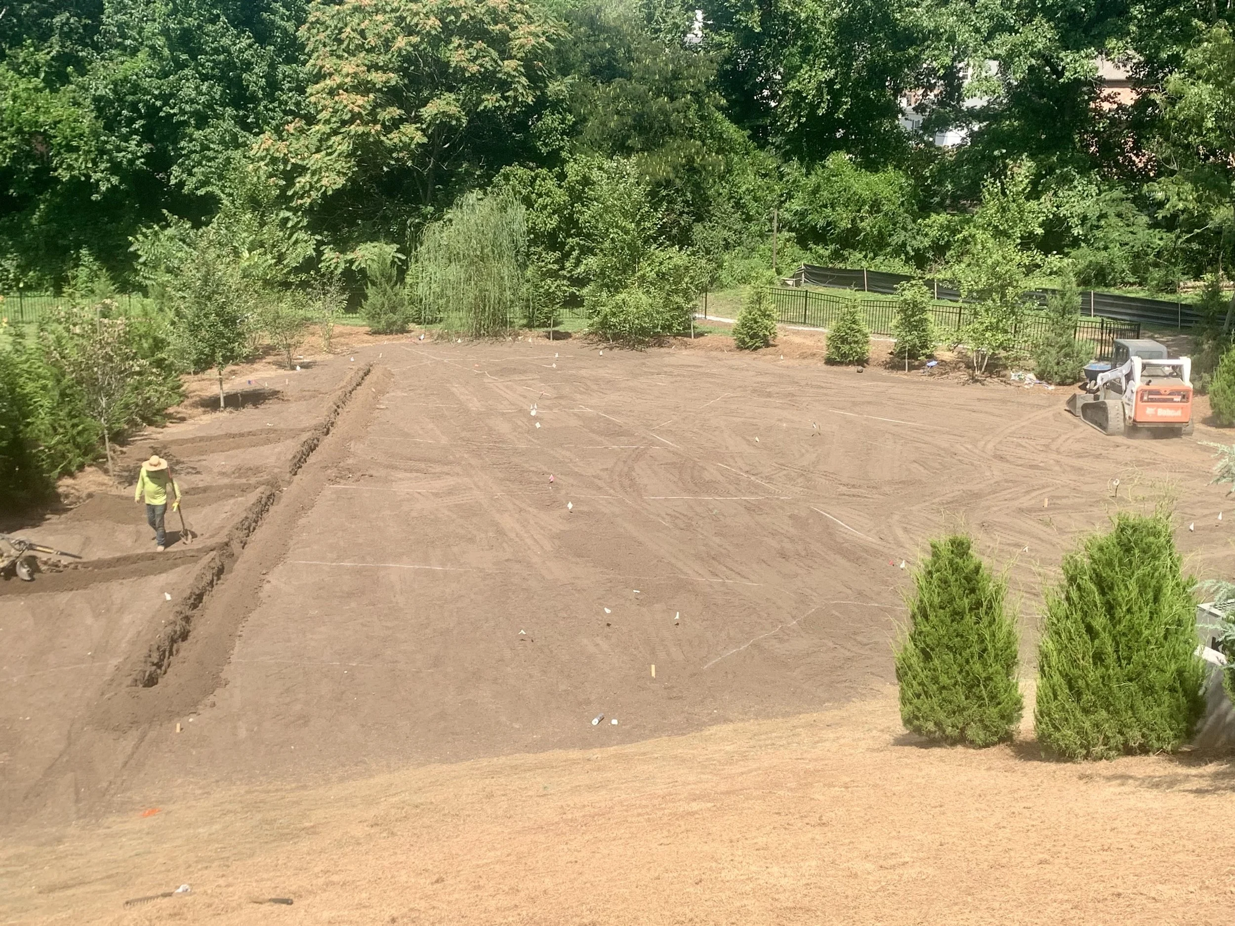 A park or garden area with freshly tilled soil and small trees, with a person wearing a yellow outfit and straw hat working in the site, overgrown trees in the background, and construction equipment and fencing present.